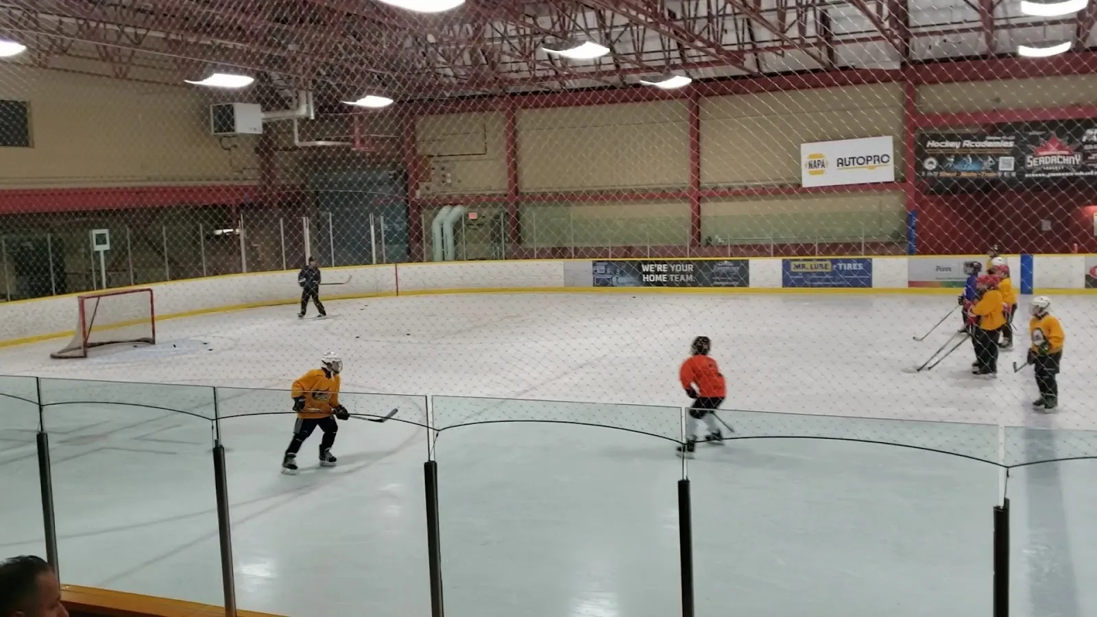 A game under a dark red-trussed roof, players spread across the sheet with netting along the near boards and a Canadian flag on the far wall. — Callingwood Twin Arena