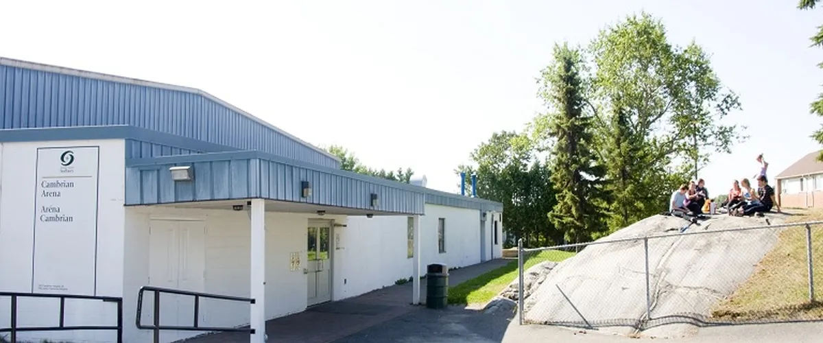 The arena’s low white entrance building and bilingual Cambrian Arena sign, with trees and a rock outcrop beside it. — Cambrian Arena