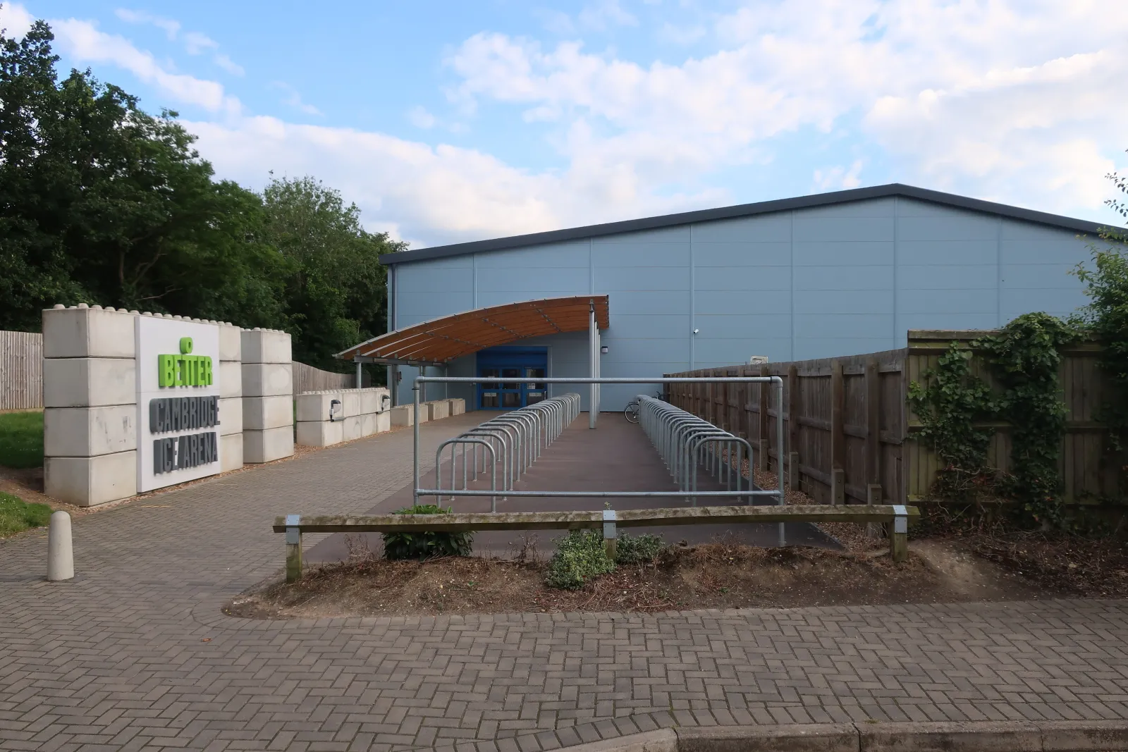 The entrance to Cambridge Ice Arena, a grey clad hall with a curved timber canopy over the door, a Better sign by the path and bike racks along the ramp. — Cambridge Ice Arena