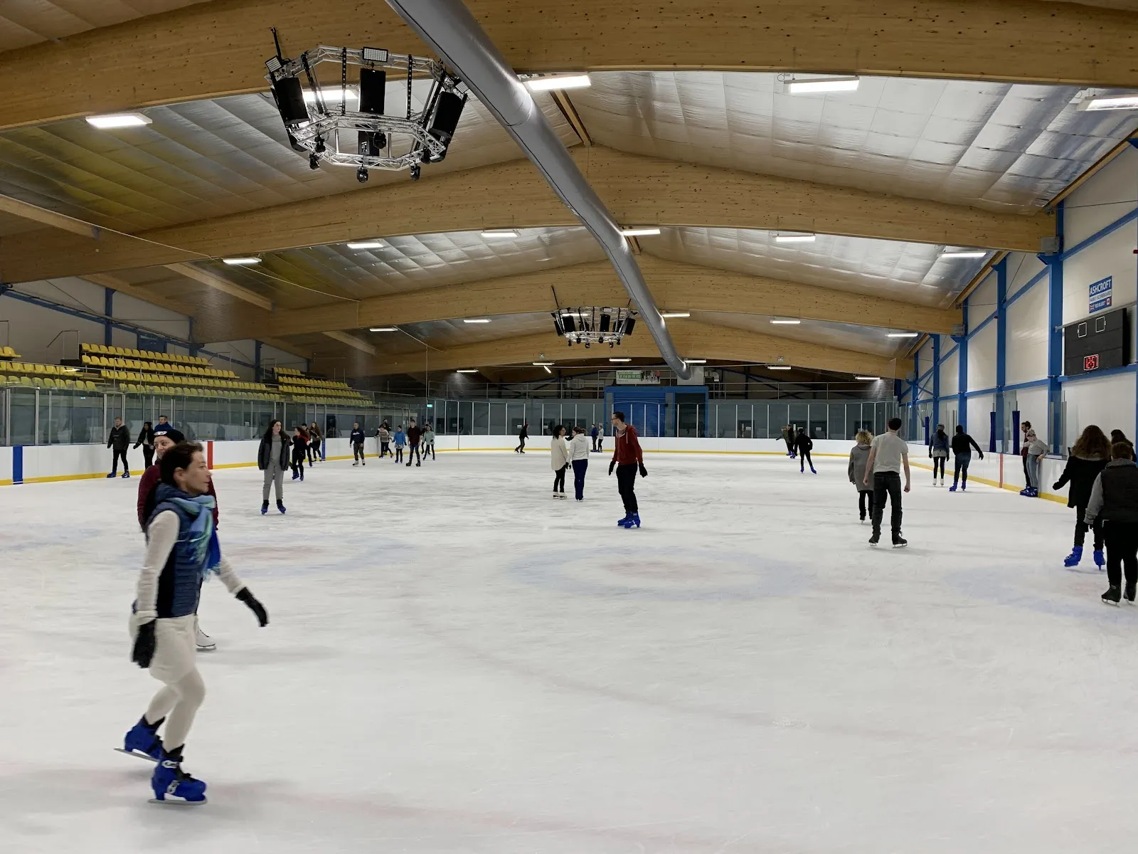A public session on the ice at Cambridge Ice Arena, skaters spread across the rink under the curved timber roof with blue steel columns along the boards. — Cambridge Ice Arena