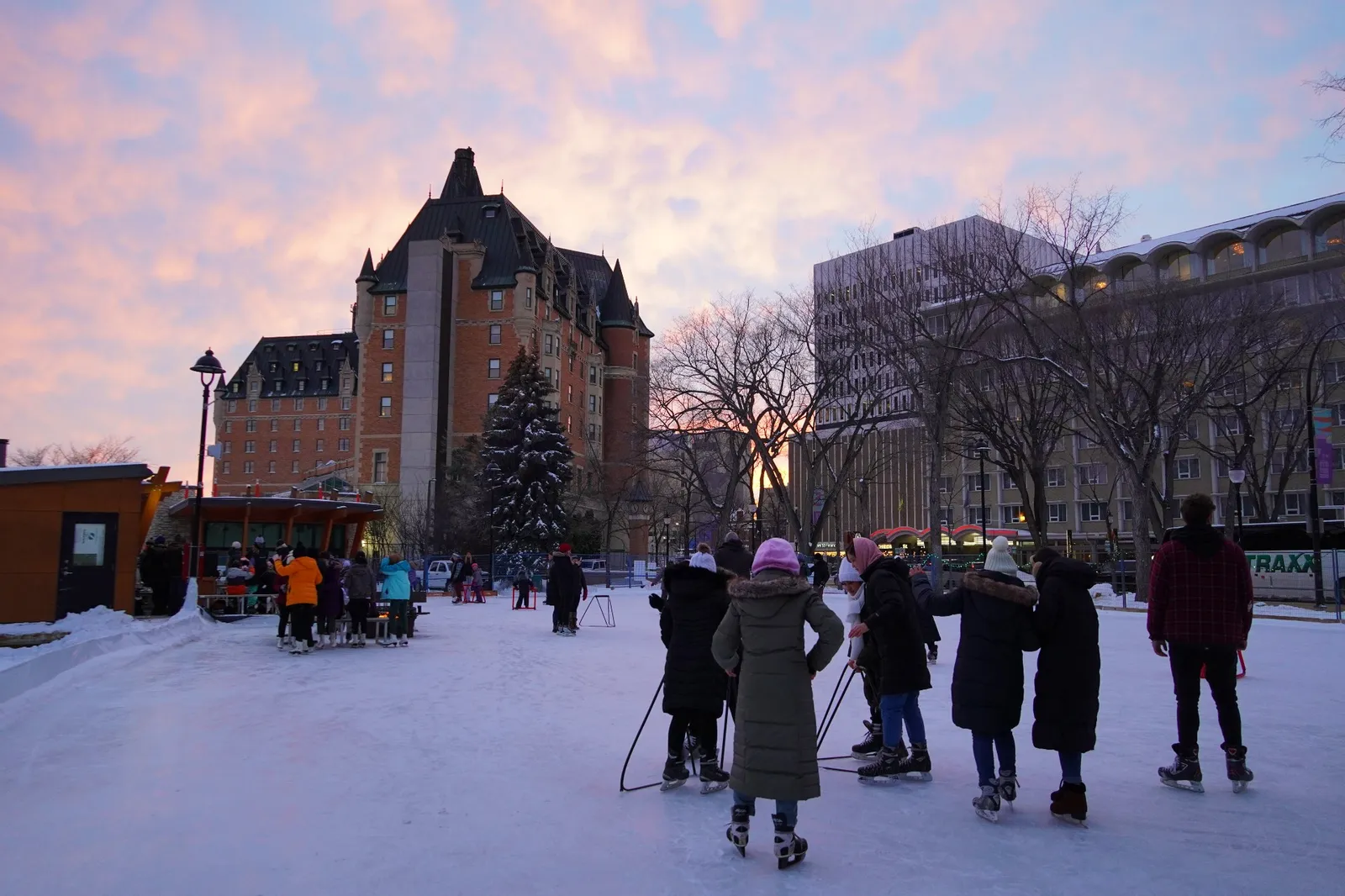 Skaters on the Cameco Meewasin Skating Rink at Nutrien Plaza in Saskatoon at dusk, with the Delta Bessborough hotel rising behind the trees. — Cameco Meewasin Skating Rink @Nutrien Plaza