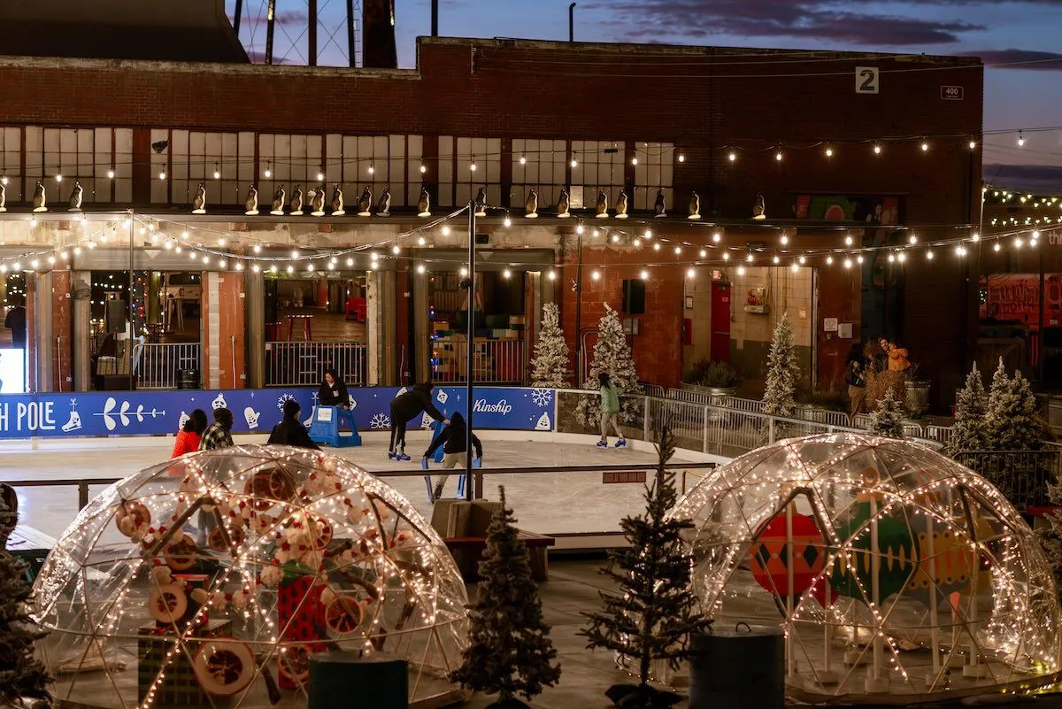 The Camp North Pole rink with igloo domes and the brick Camp North End building at dusk (Camp North End website) — Camp North Pole Ice Skating