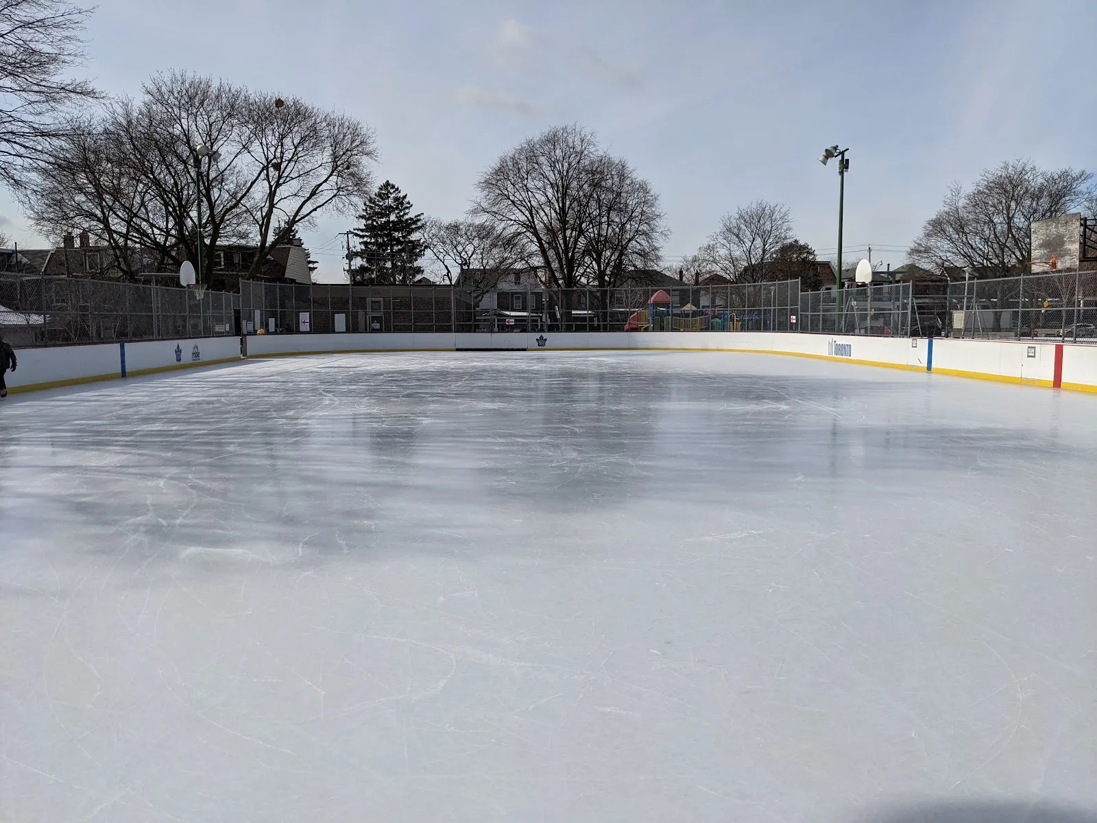La pista vacía de Campbell Avenue Playground, con bardas blancas, cercas y árboles sin hojas al fondo, sin personas a la vista — Campbell Avenue Playground Outdoor Ice Rink