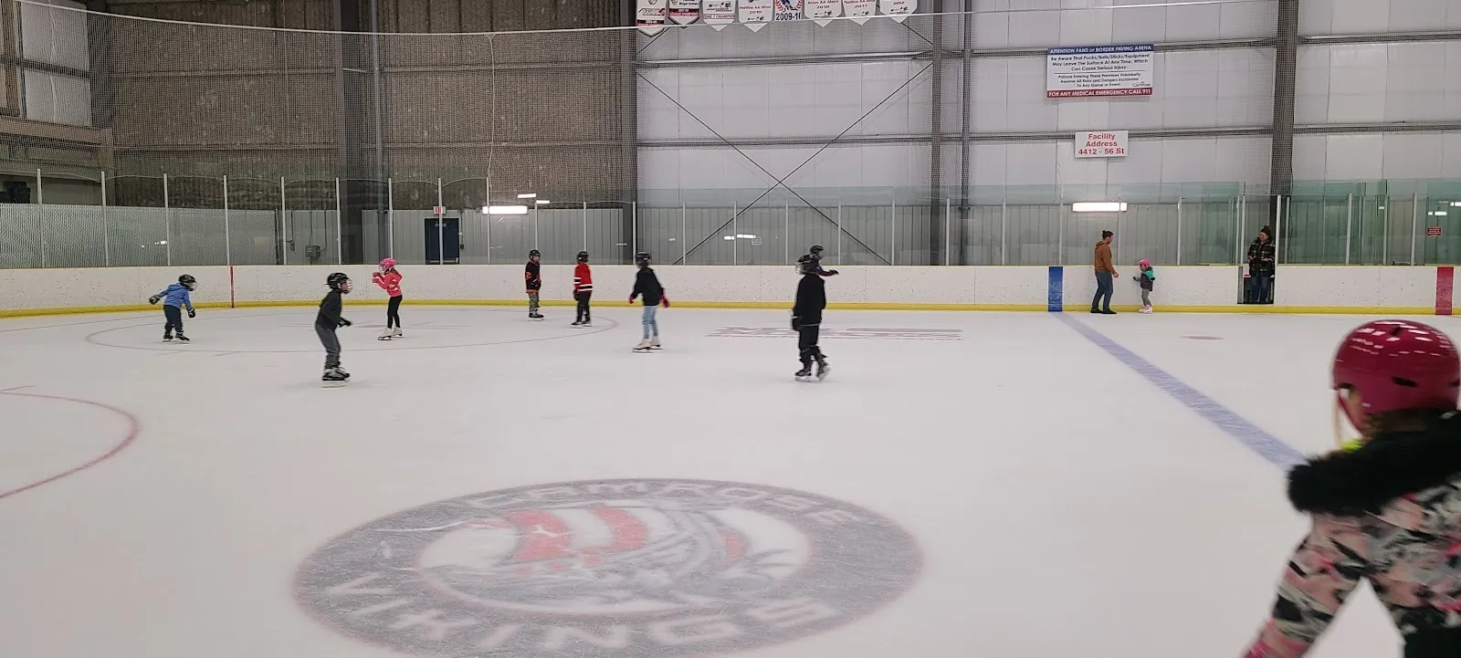 Unos cuantos patinadores repartidos por una pista amplia con un logotipo de los Vikings en el centro, con anuncios de patrocinadores y una pared gris al fondo detrás de ellos. — Camrose Recreation Centre