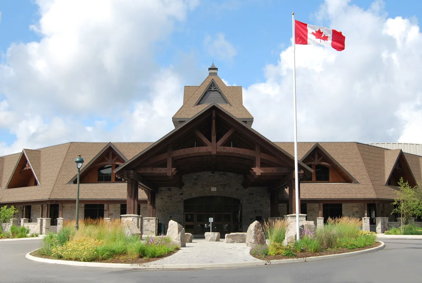 La entrada principal con estructura de madera del Canada Summit Centre, con base de piedra, techo a dos aguas y la bandera de Canadá. — Canada Summit Centre (Huntsville)