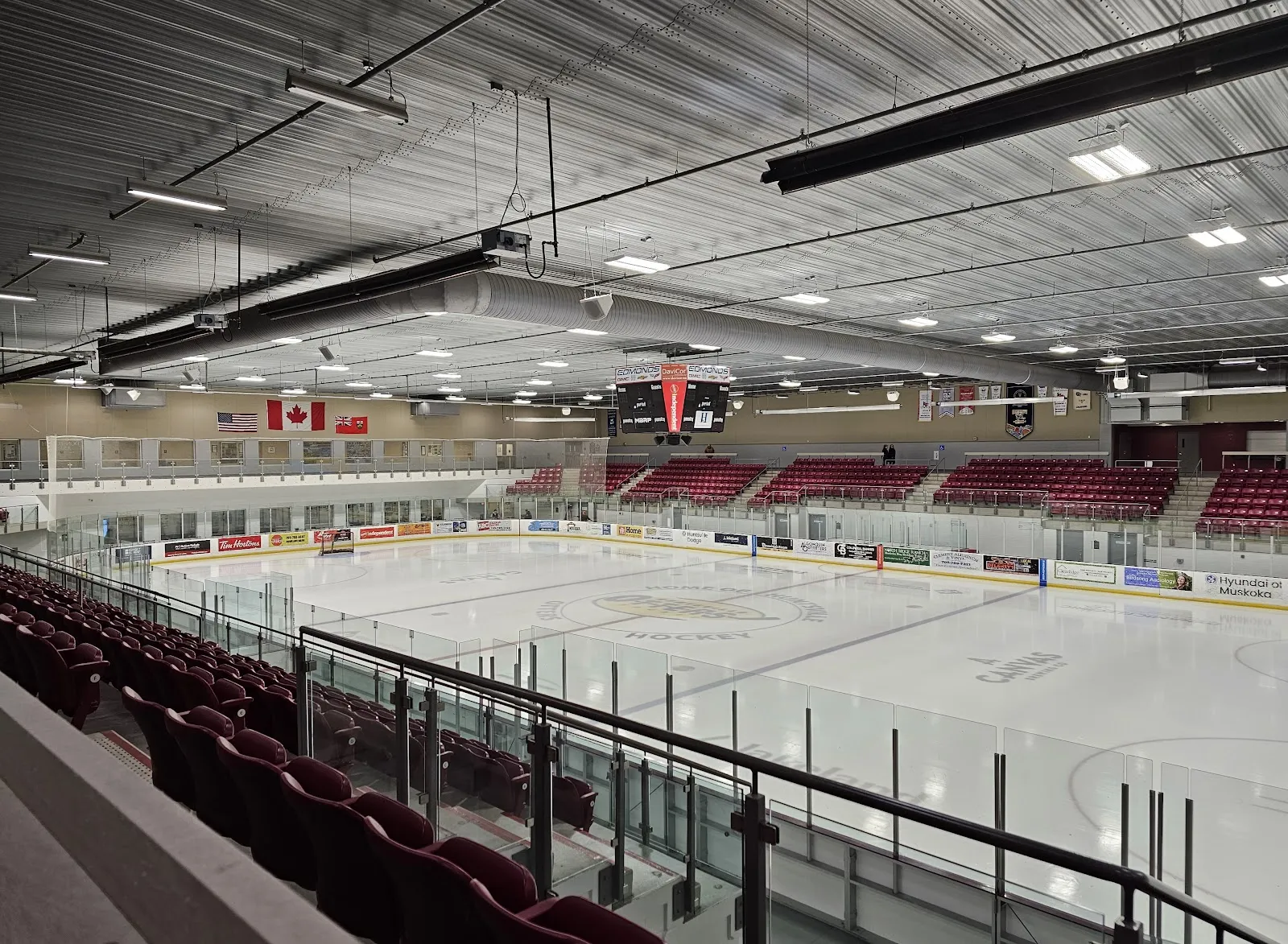 El hielo vacío de la Don Lough Arena desde los asientos superiores, con asientos rojos a ambos lados. — Canada Summit Centre (Huntsville)