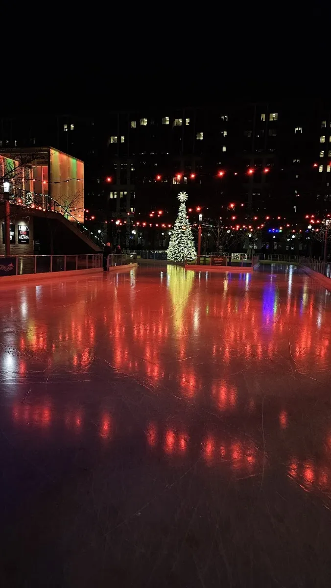 The empty Canal Park rink at night glowing red from the lights, with a lit tree and apartment blocks behind. — Canal Park Ice Rink