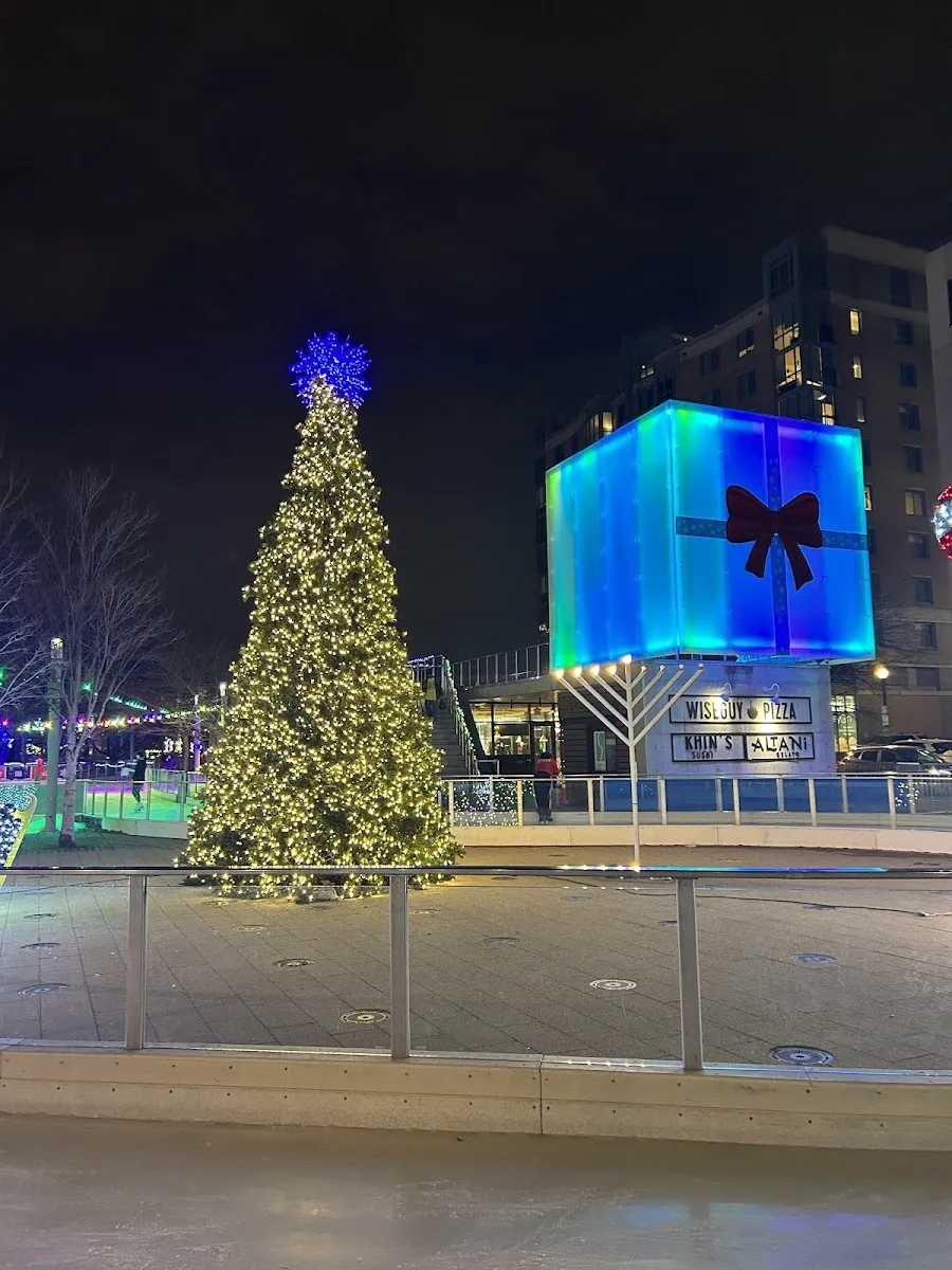 The Canal Park rink beside its lit Christmas tree and a blue glowing cube sculpture in the evening. — Canal Park Ice Rink