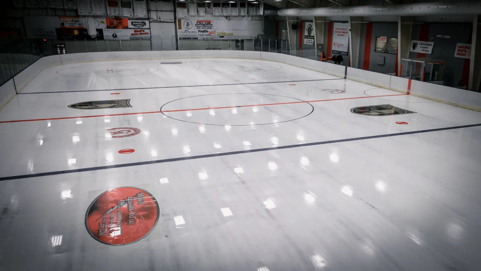 Interior view of the full Ingle Ice Arena ice sheet from ice level, with rink boards, sponsor logos on the ice, and "Canandaigua Knights Hockey" / "Canandaigua Beavers Hockey" team banners on the far wall. — Greater Canandaigua Civic Center