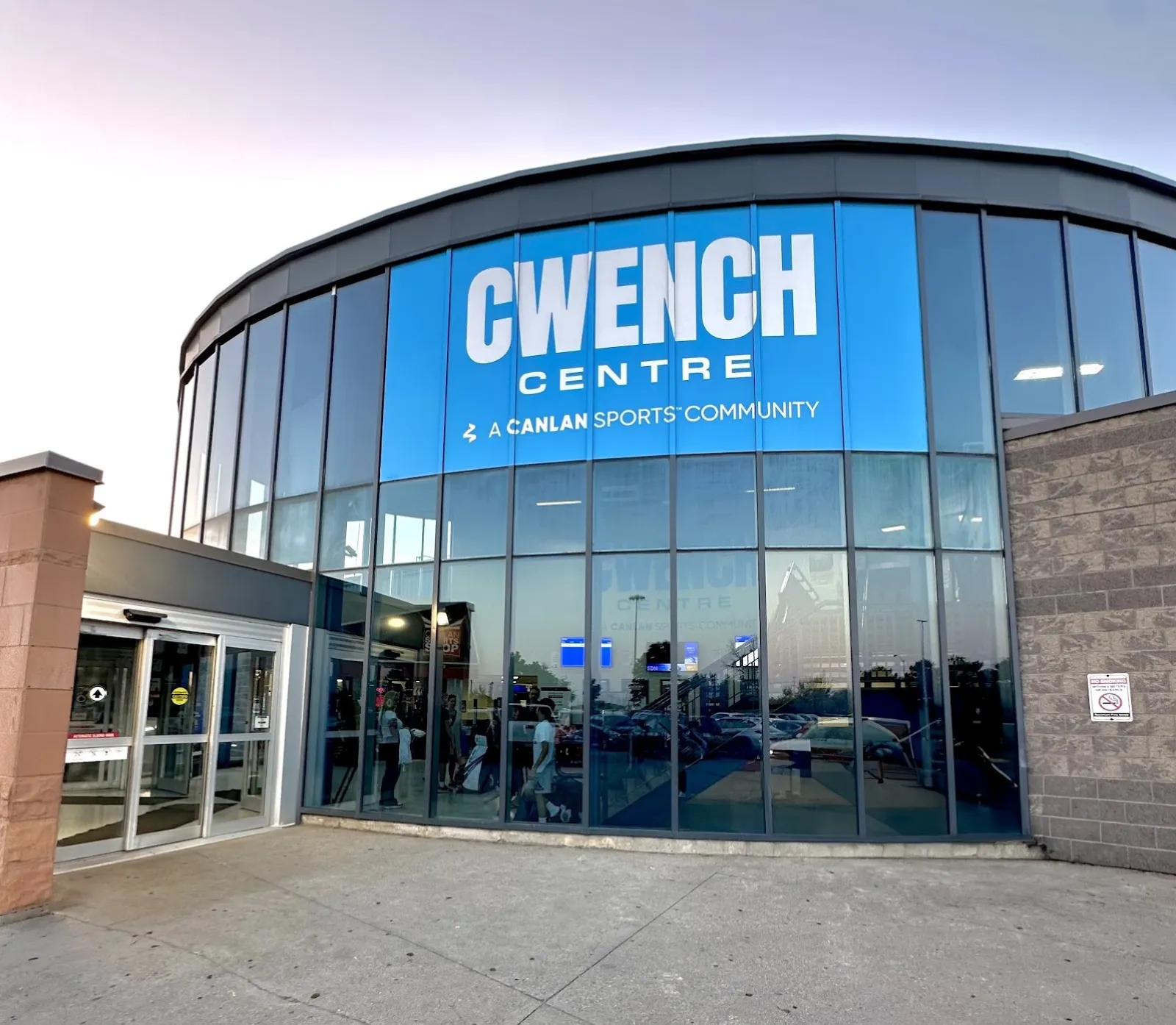 Curved glass entrance of the building at dusk, with large blue CWENCH CENTRE / A CANLAN SPORTS COMMUNITY signage above automatic sliding doors and a gray brick corner — CWENCH Centre - Etobicoke