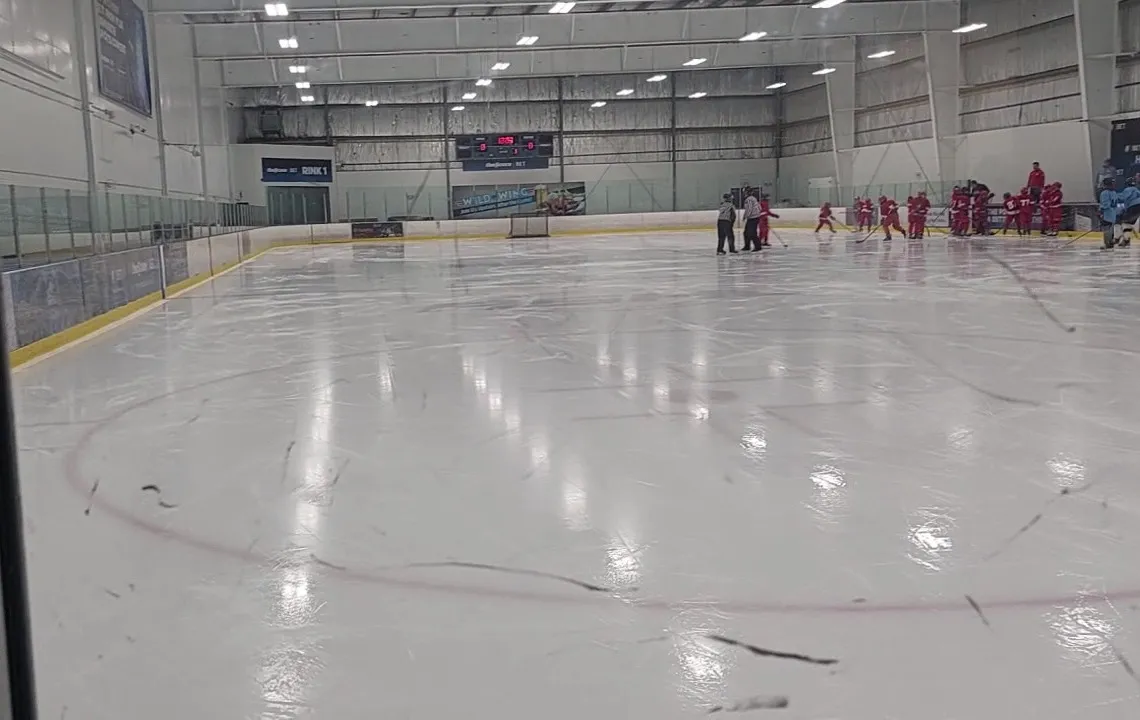 Rink 1 at the CWENCH Centre in Etobicoke from behind the glass, with a team gathered at the far end under the scoreboard — CWENCH Centre - Etobicoke