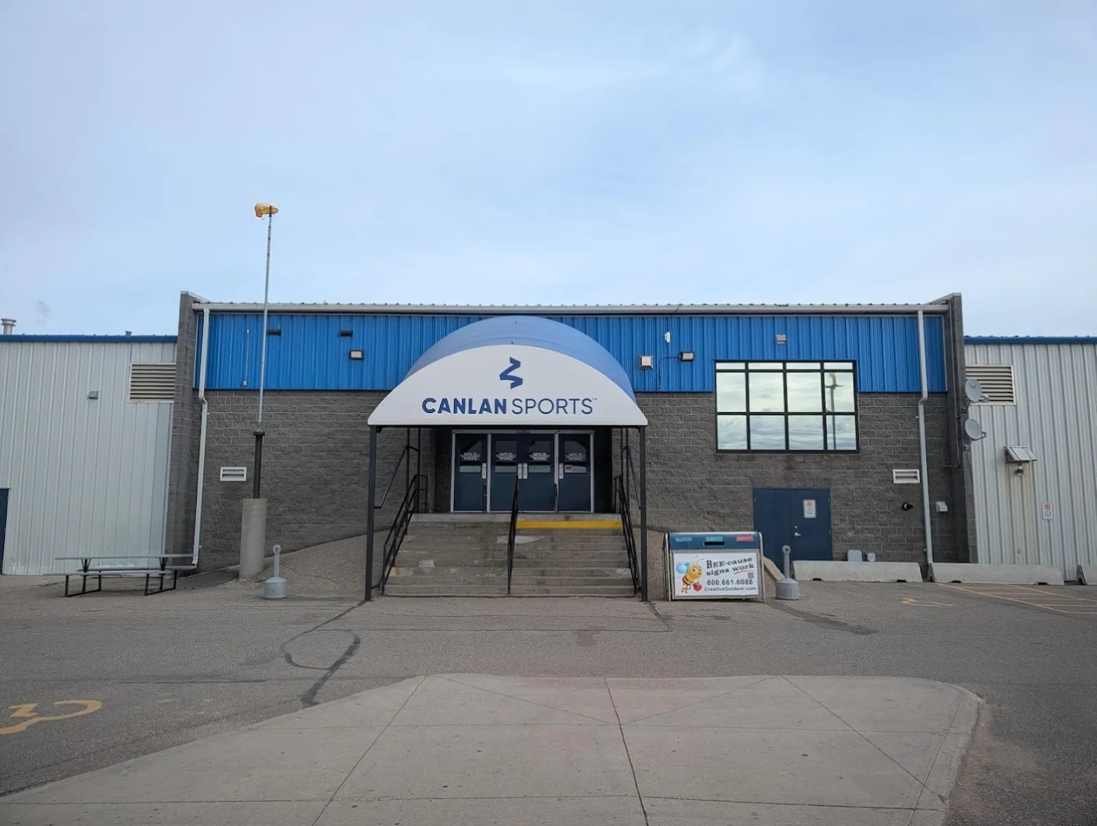 The entrance from the car park: a low grey brick and blue-clad building with a Canlan Sports canopy over the doors, a flight of steps and a wheelchair parking space painted on the asphalt. — Canlan Sports Saskatoon South (Jemini)