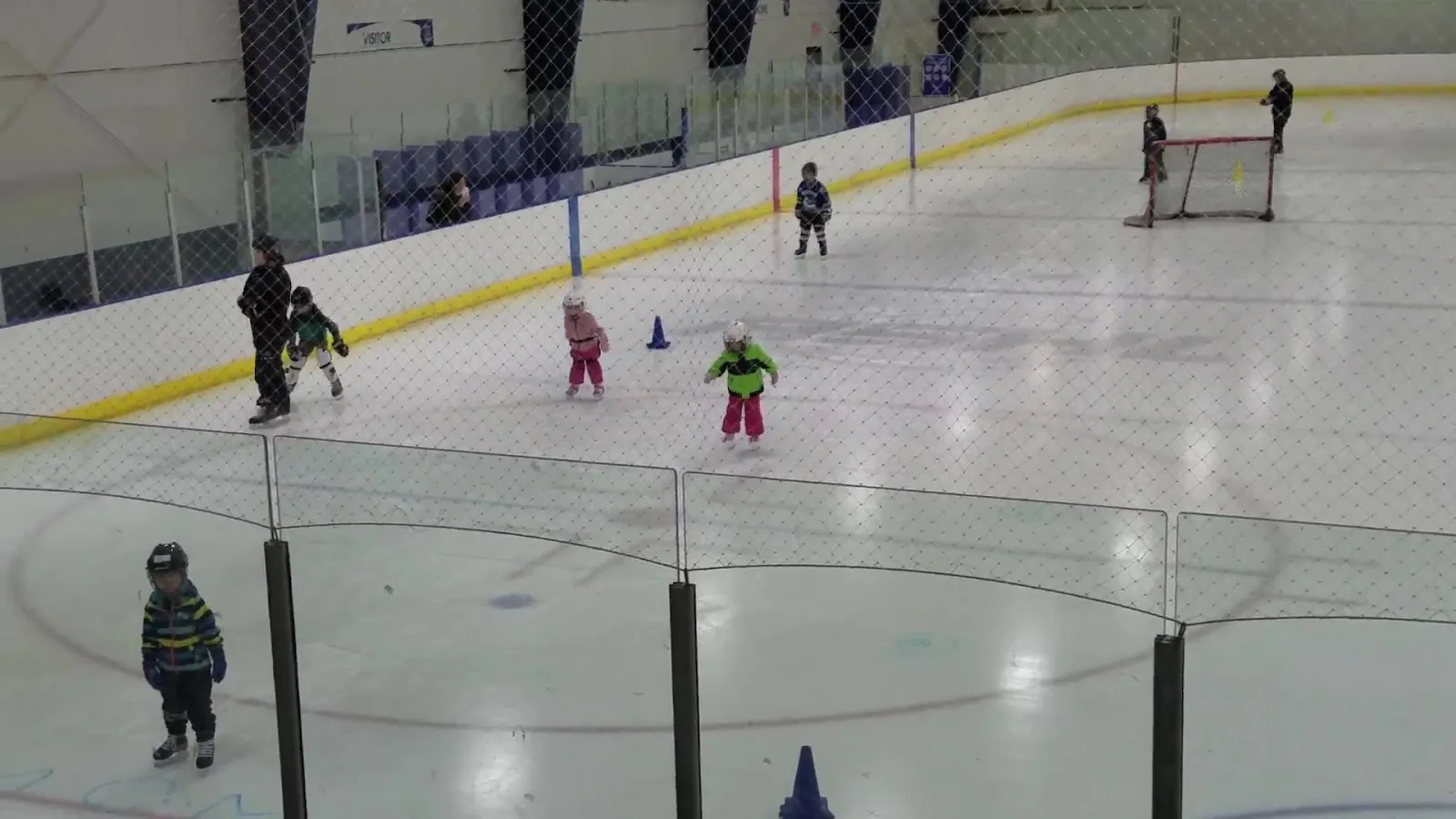 A skating session on the ice at Canlan Sports Saskatoon South: young skaters spread across the sheet with training cones, seen through the glass and netting with the goal and boards behind them. — Canlan Sports Saskatoon South (Jemini)