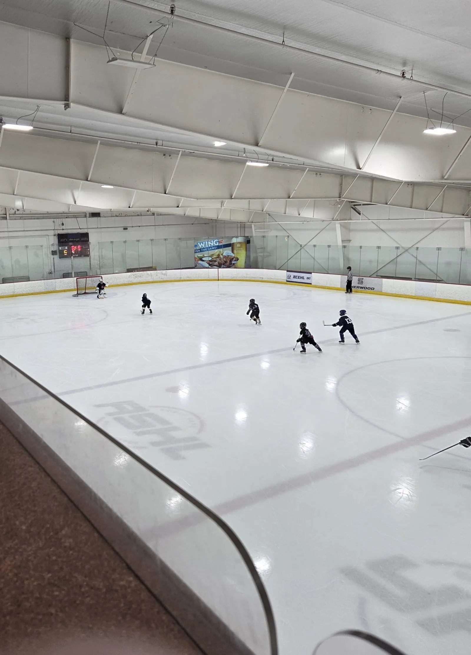 Wide view of one NHL-sized ice sheet from the stands, boards and glass around the rink, a scoreboard and a Wild Wing ad board on the far boards, with youth players mid-drill. — Canlan Sports Langley Twin Rinks