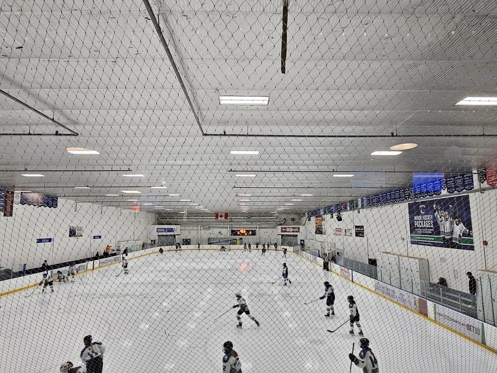 Una vista amplia desde las gradas de una pista con patinadores y jugadores repartidos por el hielo bajo un techo de acero con banderines en la pared del fondo. — Canlan Sports North Shore