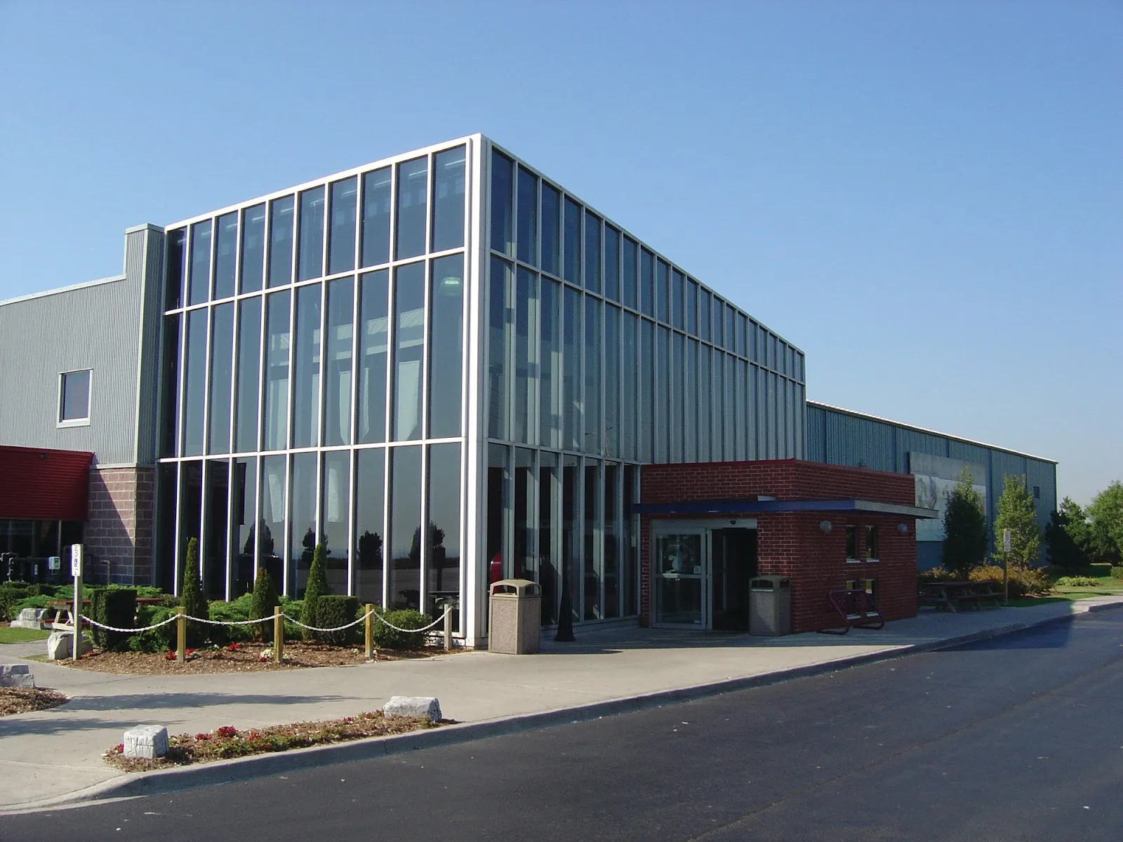 The Canlan Sports Oshawa building on a sunny day: a two-storey glass entrance block against the grey arena walls, with a red-brick vestibule and landscaped front walk. — Canlan Sports Oshawa