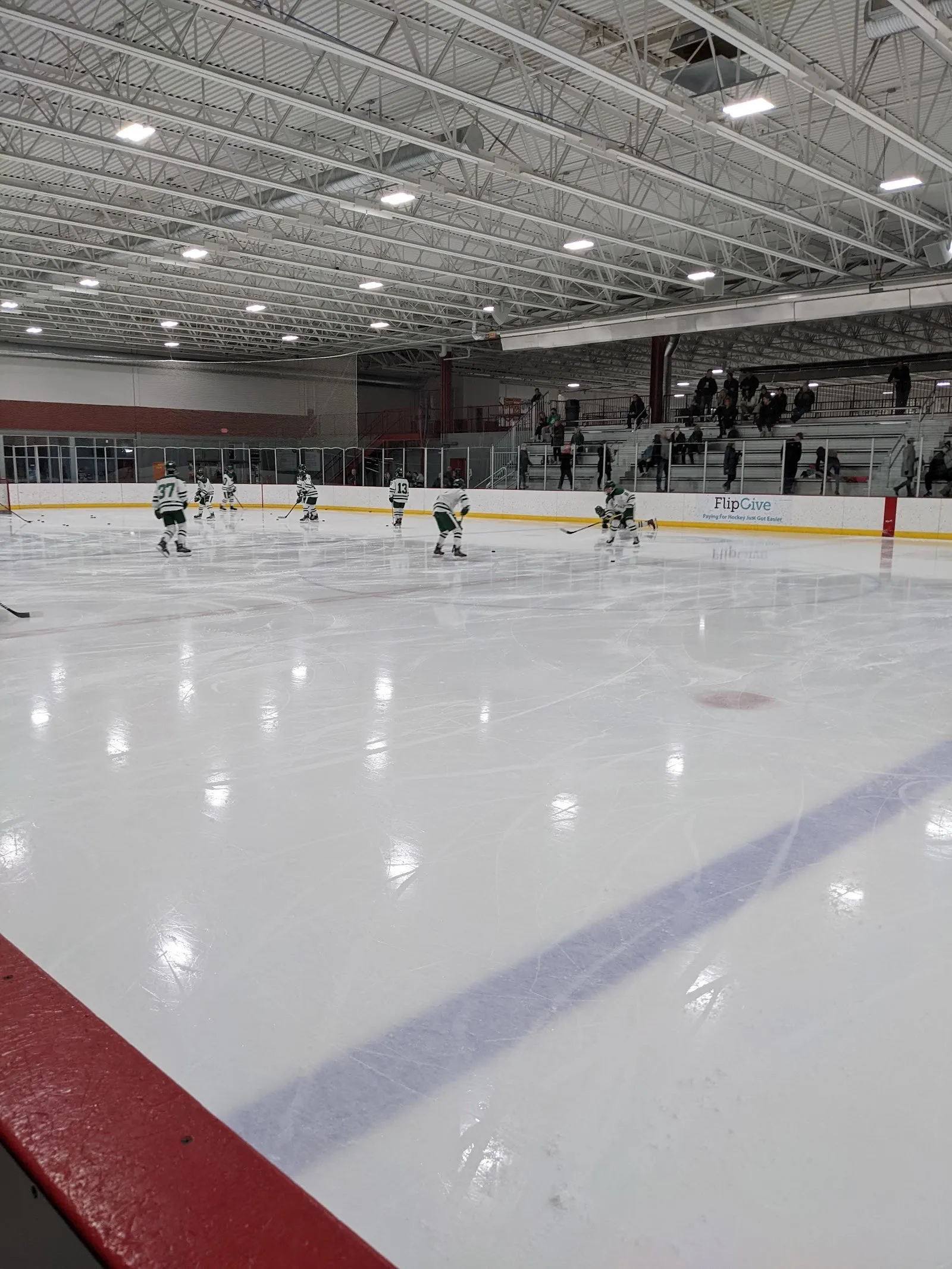 A sheet at Canlan Sports Romeoville from the bench door, with a team warming up at the far end below the stands — Canlan Sports Romeoville