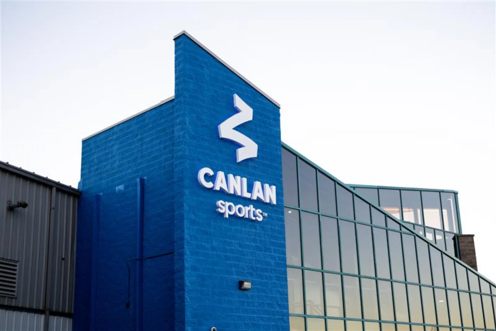 Corner of the blue-brick building with a lit "CANLAN sports" wordmark and logo sign, glass-curtain entrance wing to the right. — Canlan Sports Scarborough