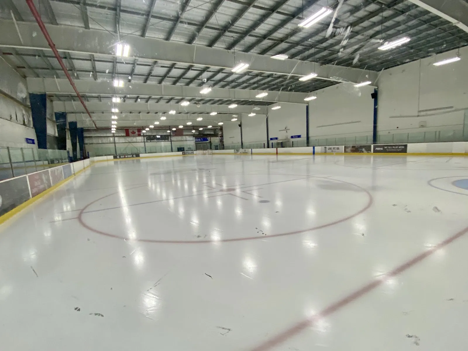 Empty ice sheet on Rink 1 shot rinkside toward the far boards, red and blue faceoff/centre lines visible, Canadian and American flags hung above the far glass, steel-truss ceiling overhead, no people on the ice. — Canlan Sports Scarborough