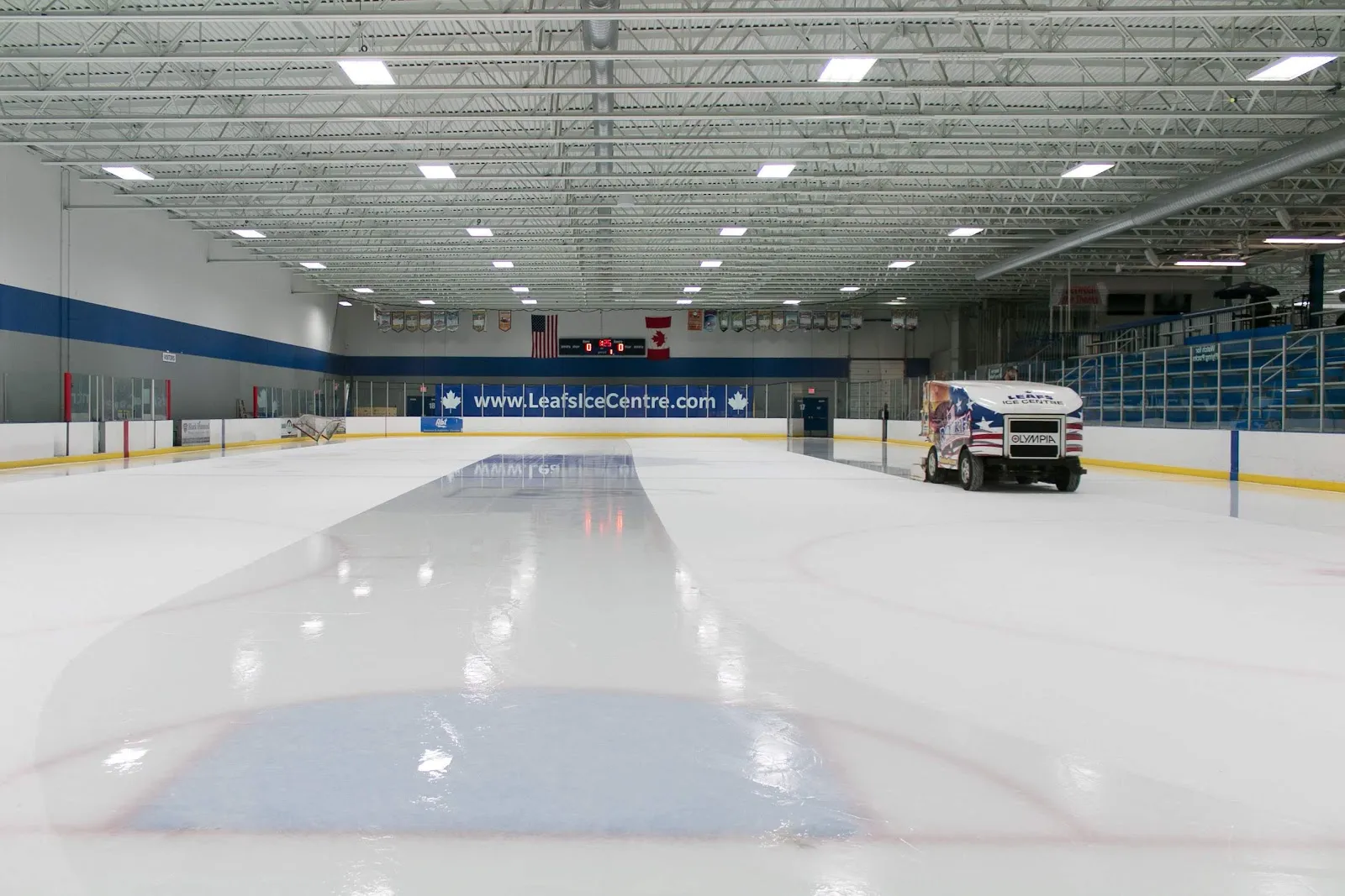 Empty ice sheet under exposed steel roof trusses, dasher boards reading "www.LeafsIceCentre.com" with US and Canadian flags above the scoreboard, and a Zamboni parked at rink-side — Canlan Sports West Dundee