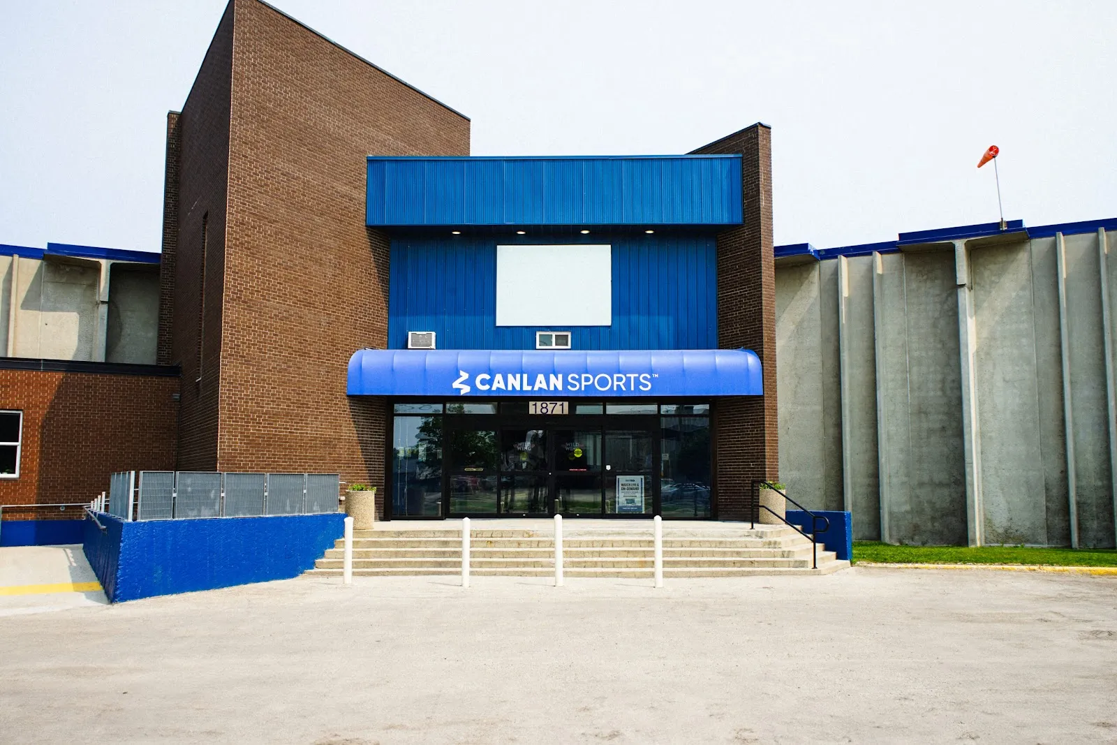 La fachada del edificio desde el estacionamiento: un toldo azul con el letrero Canlan Sports sobre las puertas de cristal, el número de la calle encima, paredes de ladrillo y hormigón y una escalinata. — Canlan Sports Winnipeg