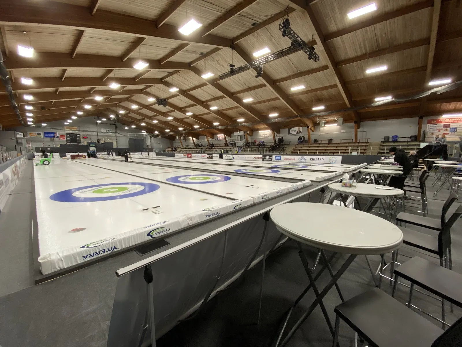 Una pista de hielo con anillos azules de curling pintados, con mesas y sillas junto al cristal para un evento, bajo un techo de madera con focos. — Canlan Sports Winnipeg
