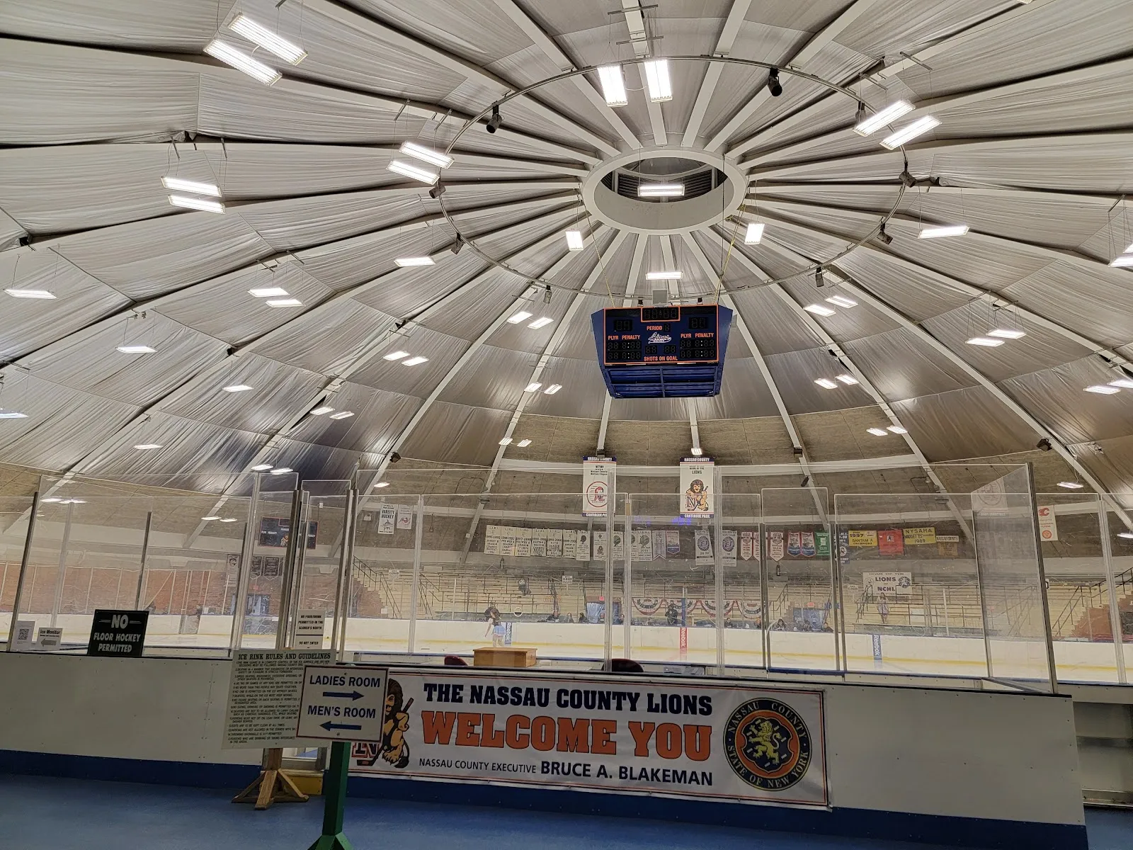 Inside the domed Cantiague Park ice rink with the rink behind the boards, the Nassau County banner and the scoreboard under the roof — Cantiague Park Ice Rink