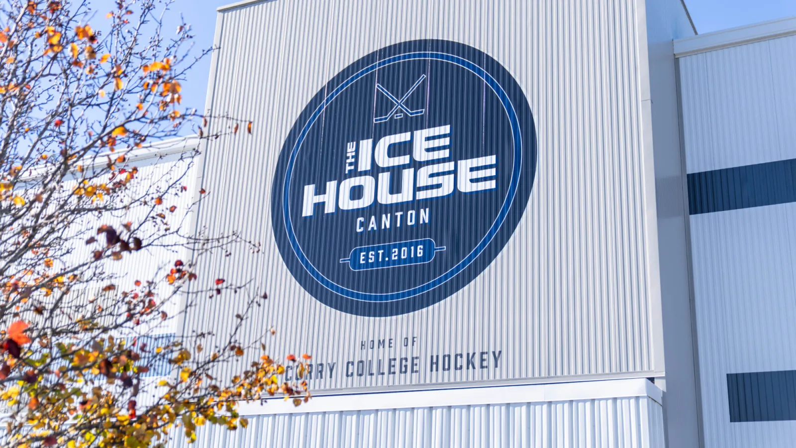 The metal-clad side of the building with a large round "The Ice House Canton, Est. 2016" sign above the words "Home of Curry College Hockey". — Canton Ice House