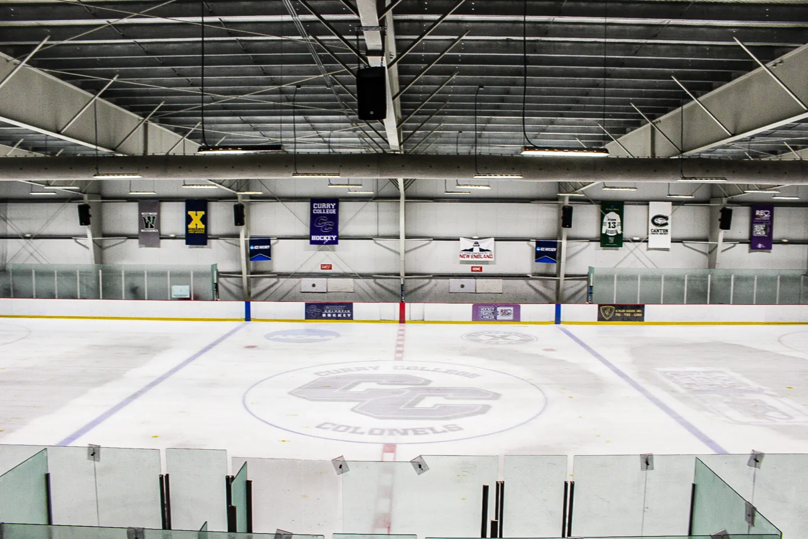 An empty rink seen over the glass from the stands, with the Curry College Colonels logo at center ice and team banners along the far wall. — Canton Ice House
