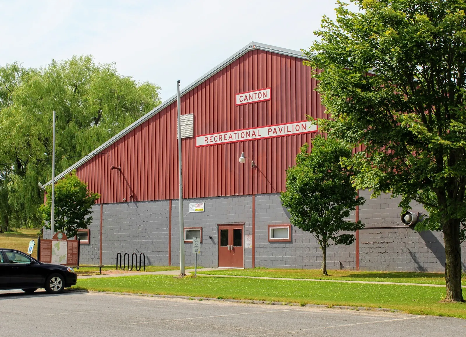 Front of the Canton Recreational Pavilion on a sunny day, red vertical-panel gable end reading CANTON RECREATIONAL PAVILION over a gray concrete-block base, double entry doors, bike rack and trees in front. — Canton Recreation Pavilion