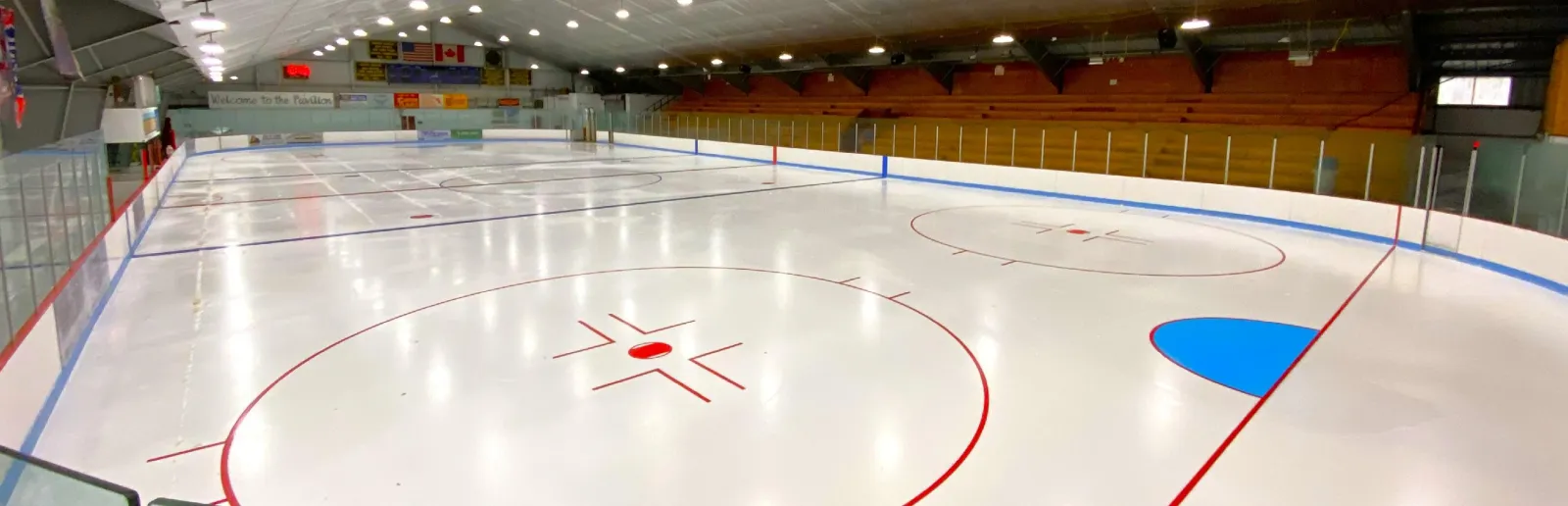 Full ice sheet inside the Pavilion, wood bleachers along the far side, a "Welcome to the Pavilion" banner and US/Canada flags on the near wall. — Canton Recreation Pavilion