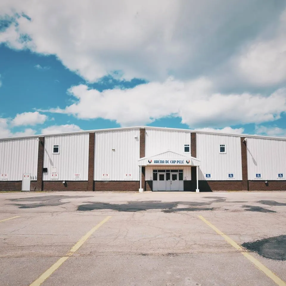 The white metal-clad Arena de Cap-Pele with brick piers and a small gabled canopy over its double doors, seen from across a wide parking lot beneath towering white clouds. — Cap-Pelé Arena