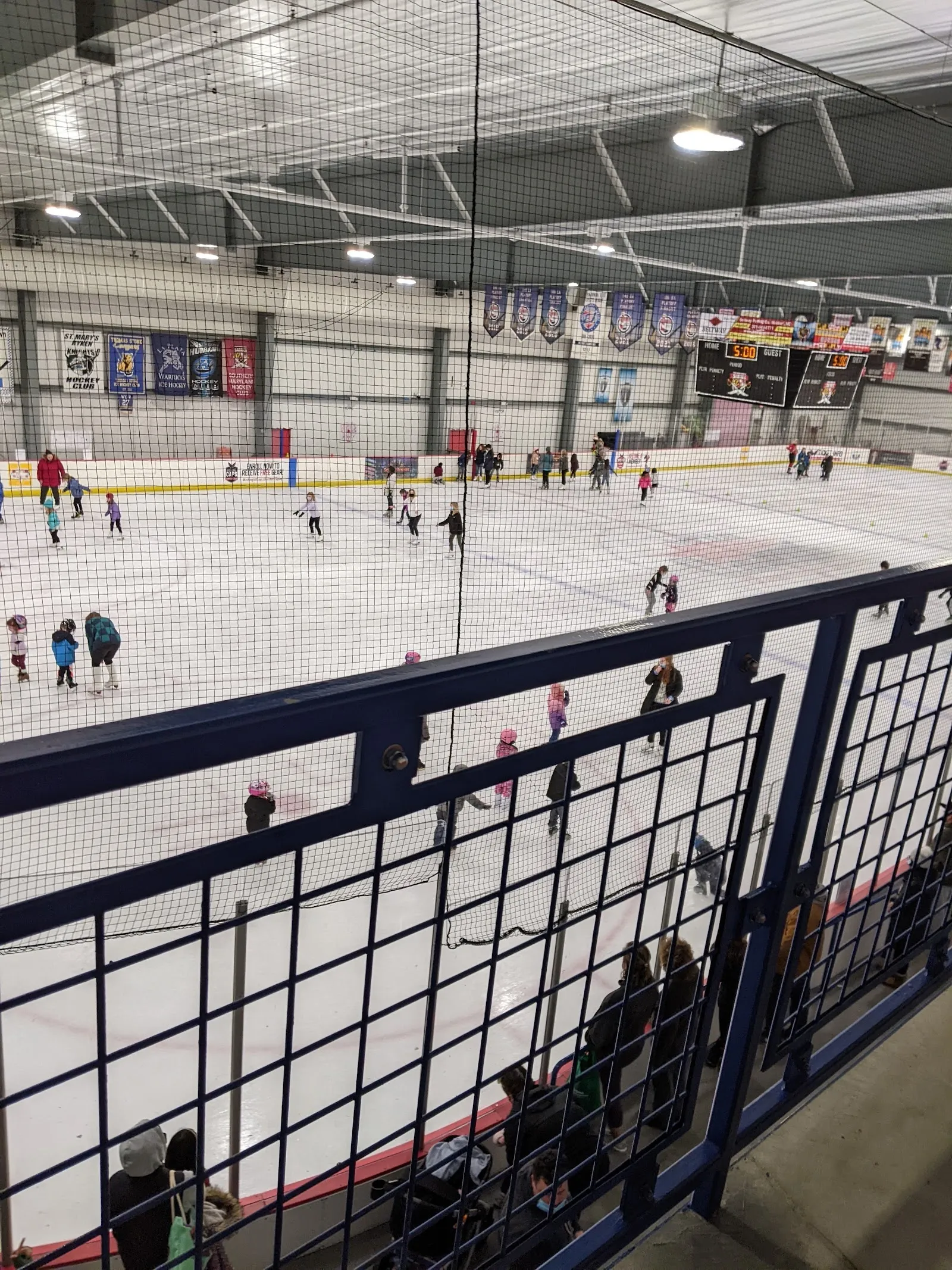 Patinadores repartidos por el hielo en el extremo opuesto de la pista, vistos por encima de un barandal metálico negro a lo largo del lado cercano. — Capital Clubhouse