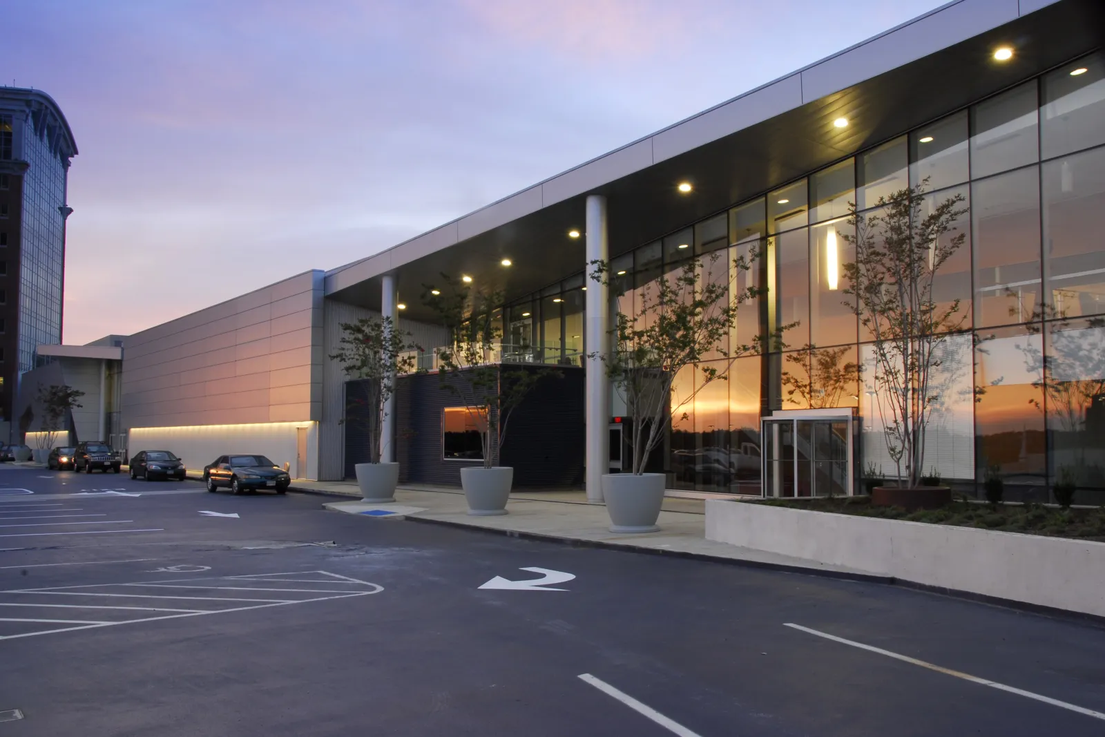Dusk exterior of the glass-and-metal-clad Iceplex atop the Ballston parking garage, with a covered entrance colonnade, planters and a parking area in front. — MedStar Capitals Iceplex