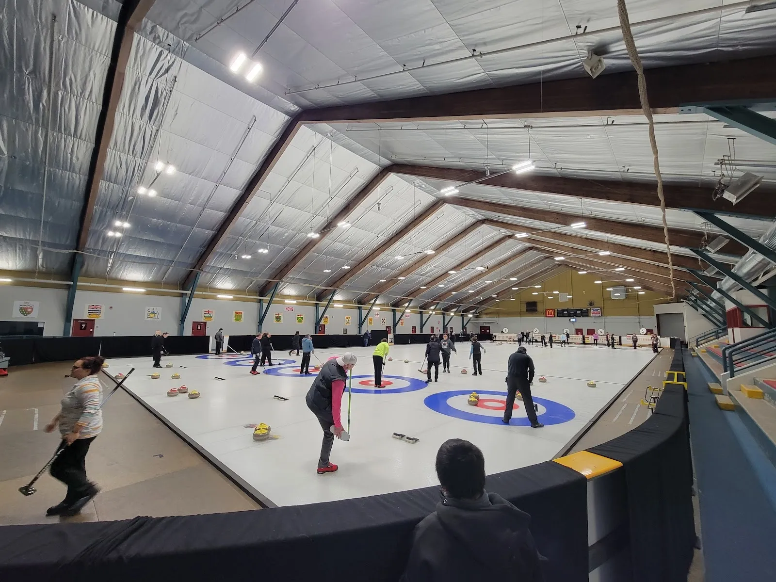 A curling session on a wide sheet under a timber-and-steel roof, people spread across the lanes around blue rings. — Capri Pizzeria Recreation Complex