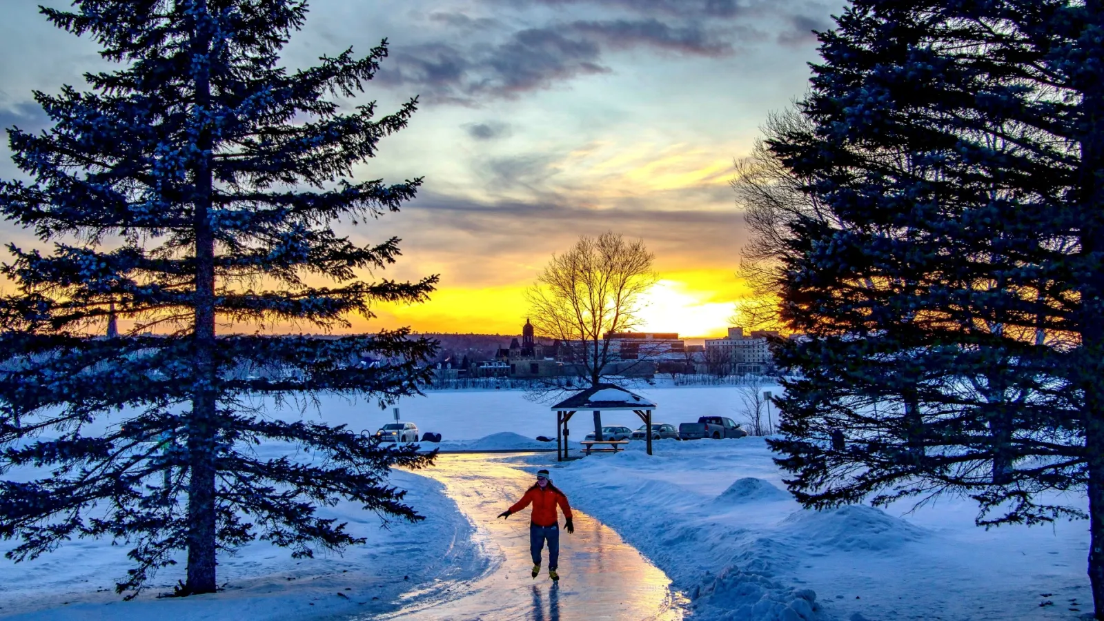 Un patinador en el sendero de hielo iluminado del circuito de patinaje de Carleton Park al atardecer, entre abetos, con un pequeño mirador y el río al fondo. — Carleton Park Natural Outdoor Rink