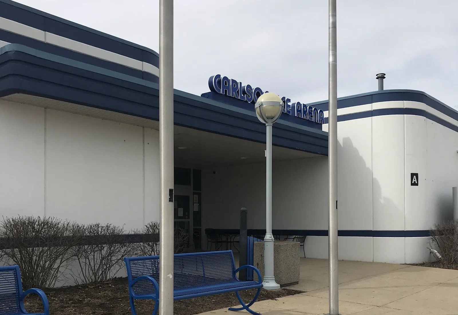 The Carlson Ice Arena entrance under its lettered canopy, a white building with rounded corners banded in navy blue, with benches and flagpoles in front — Carlson Ice Arena