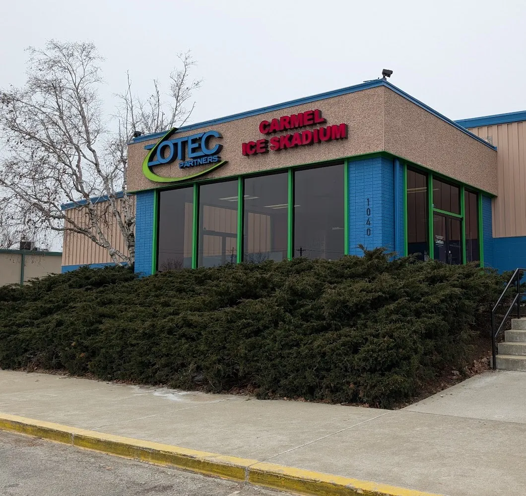 The Carmel Ice Skadium entrance at 1040, a tan block with red lettering over green-framed windows on a blue base, behind low evergreens — Carmel Ice Skadium