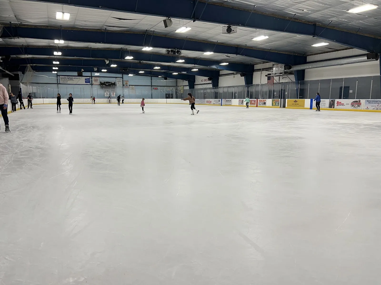 A wide sheet with skaters spread far along the boards under a dark-trussed roof, the white ice reflecting rows of ceiling lights. — Carmel Ice Skadium
