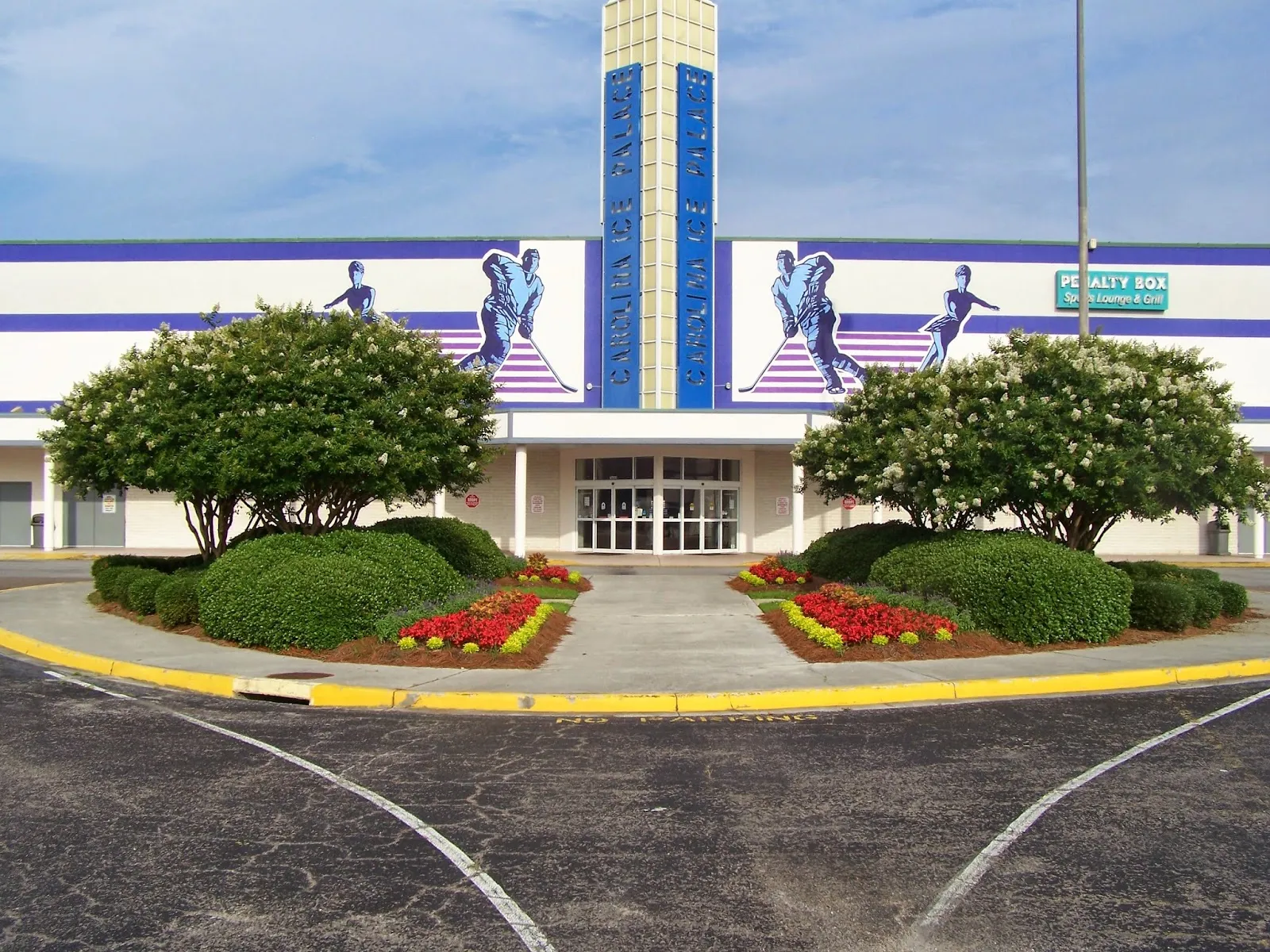 The main entrance, with a tower reading "Carolina Ice Palace" between murals of hockey players and figure skaters, and a Penalty Box sports lounge sign on the right. — Carolina Ice Palace