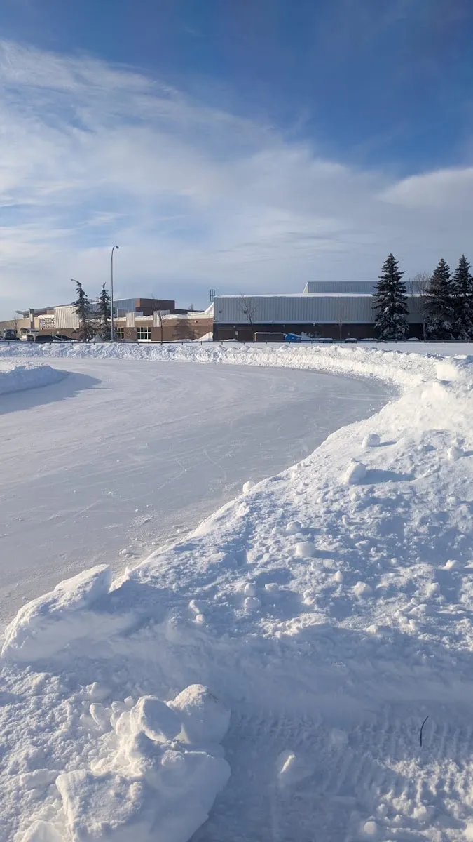 Una curva de hielo despejado en la pista al aire libre de Castle Downs en invierno, con un montículo de nieve al frente y los edificios del centro recreativo y abetos al fondo, sin personas a la vista — Castle Downs Outdoor Ice Rink