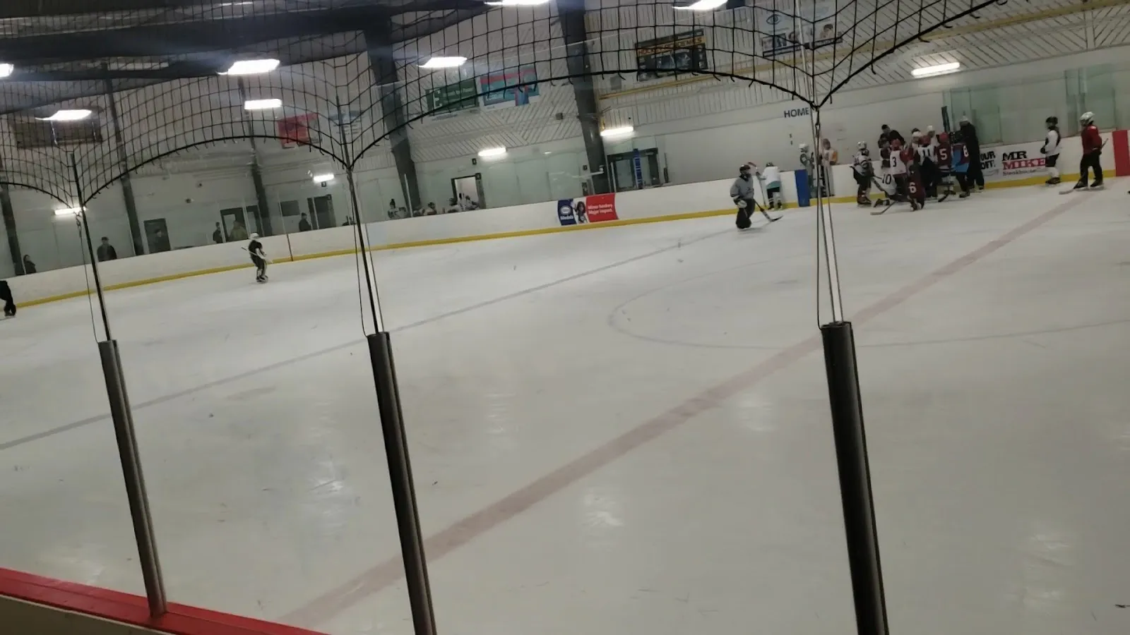 A wide view of the sheet through the netting, skaters spread along the far boards under a high roof with red lines and circles on white ice. — Castle Downs Twin Arena