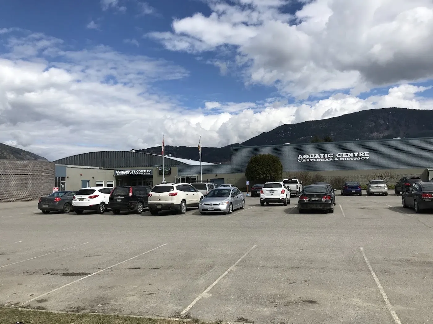 The front of the Castlegar and District complex across its car park, with the Aquatic Centre sign and the mountains behind. — Castlegar & District Community Complex