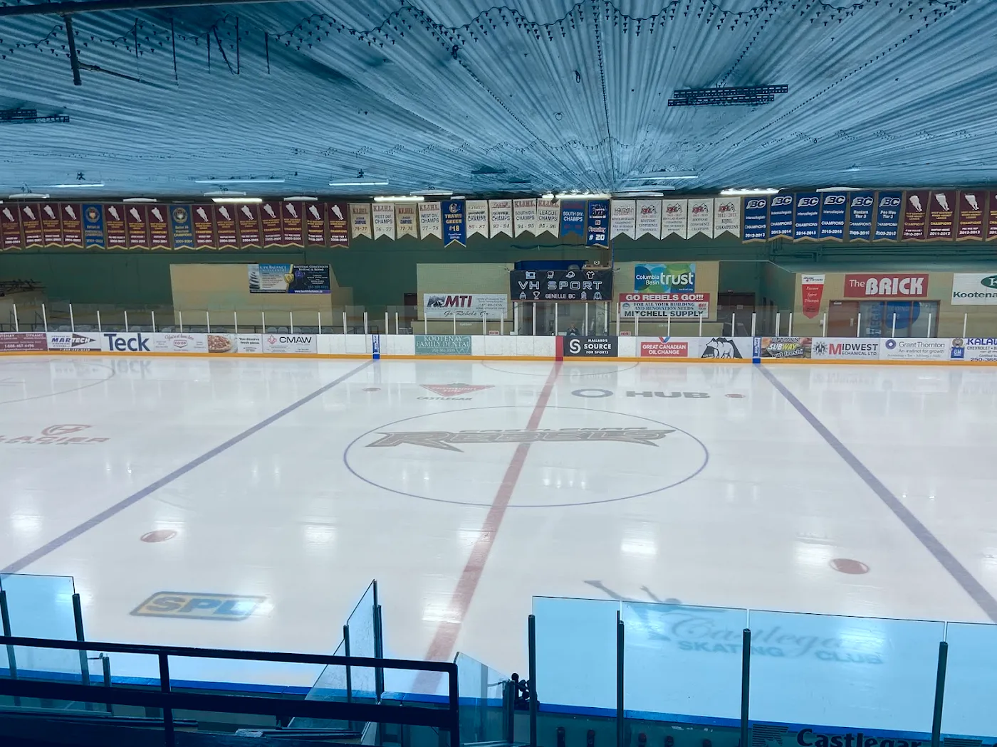 The empty ice sheet from the stands, with the centre-ice emblem, boards advertising and team banners along the far wall. — Castlegar & District Community Complex