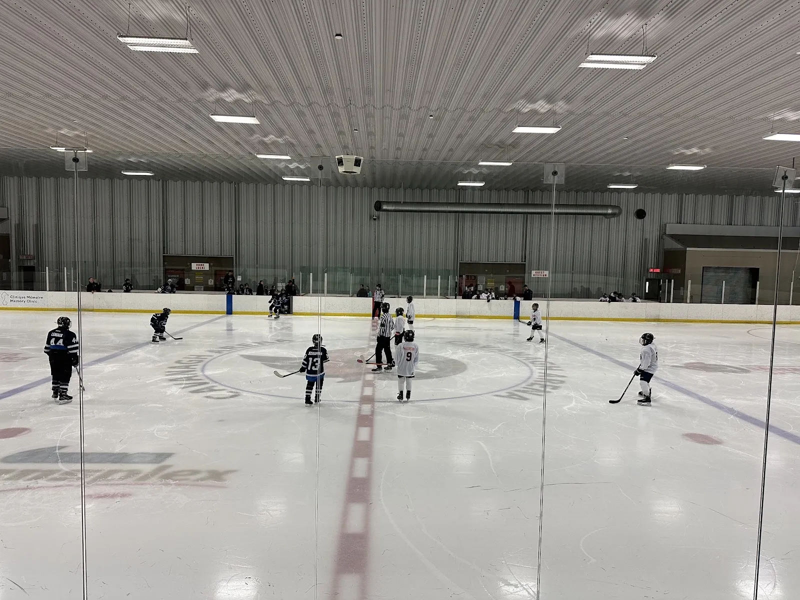 Un partido en el centro del hielo visto a través de una pista amplia, con jugadores repartidos bajo un largo techo acanalado con un gran logotipo en el centro del hielo. — Cavanagh Sensplex