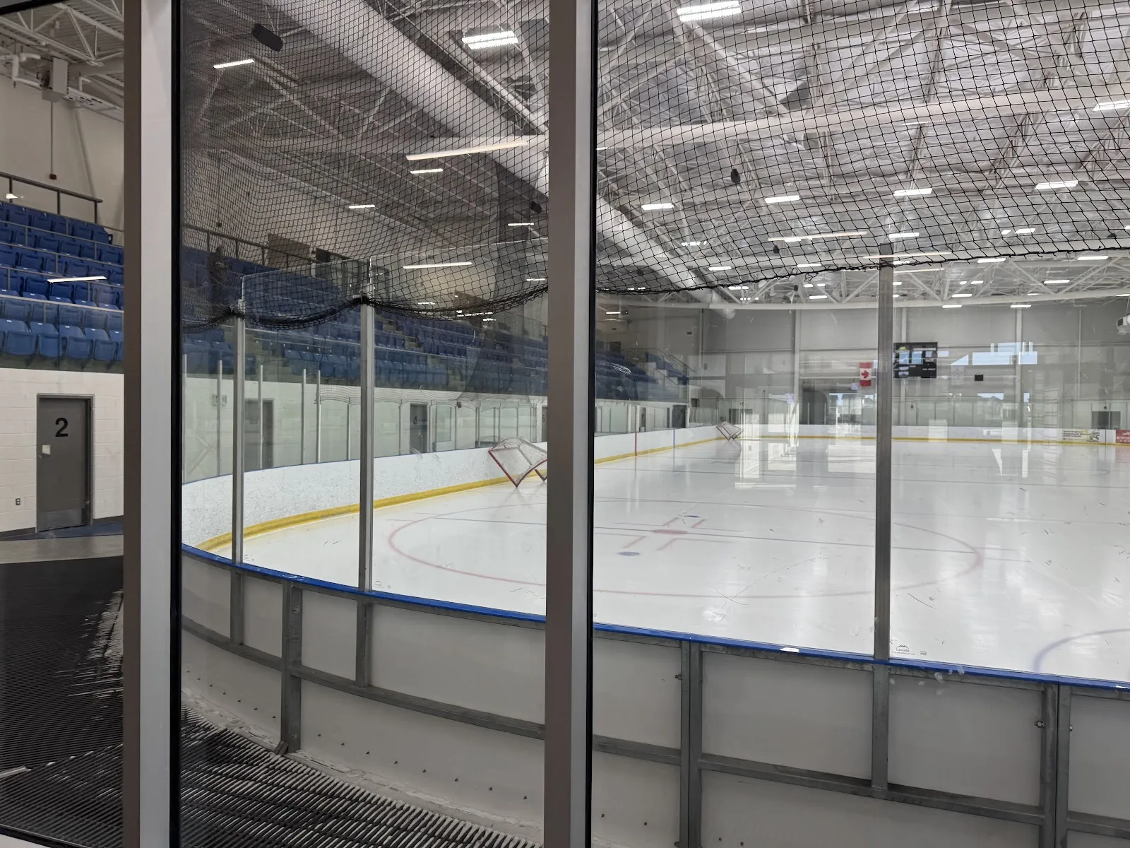 An empty NHL-size arena seen through the lobby glass, with blue seats along one side and netting above the boards. — Cavendish Farms Centre