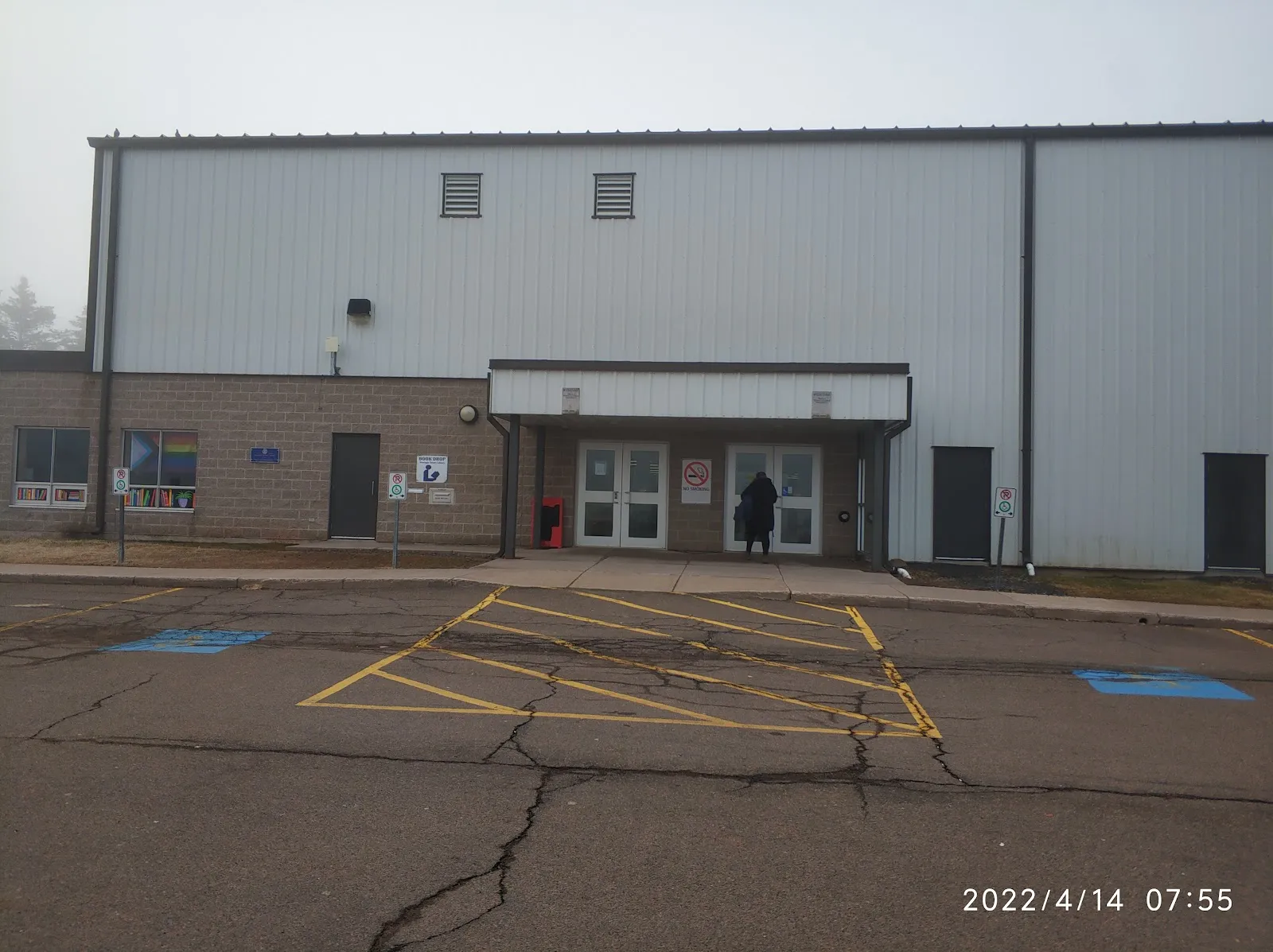 The grey metal-clad front of the Cavendish Farms Wellness Centre arena on a misty morning, with a small flat canopy over its glass double doors, a brick-faced entrance wing and accessible parking bays on the cracked asphalt in front. — Cavendish Farms Wellness Centre