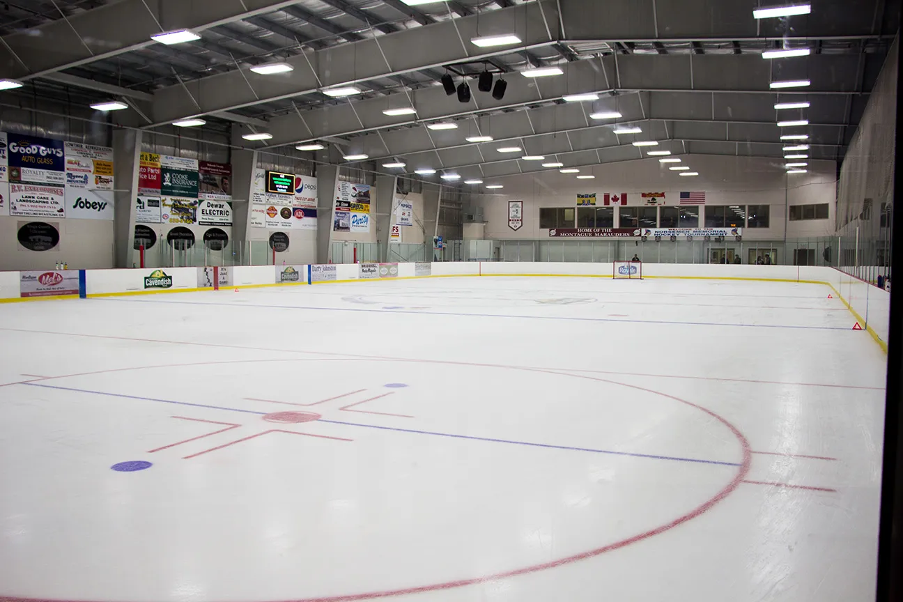 El hielo vacío visto desde la esquina, con anuncios de patrocinadores a lo largo de la pared del fondo, banderas en el extremo y un techo de acero con hileras de luces. — Cavendish Farms Wellness Centre