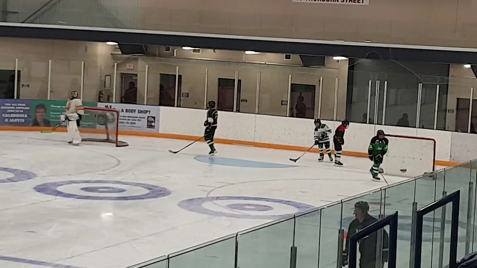 The ice surface seen from the stands through the glass, with blue circles painted on the ice, a goalie in net and a few players at the far end, and sponsor boards along the dasher. — Cayuga Memorial Arena