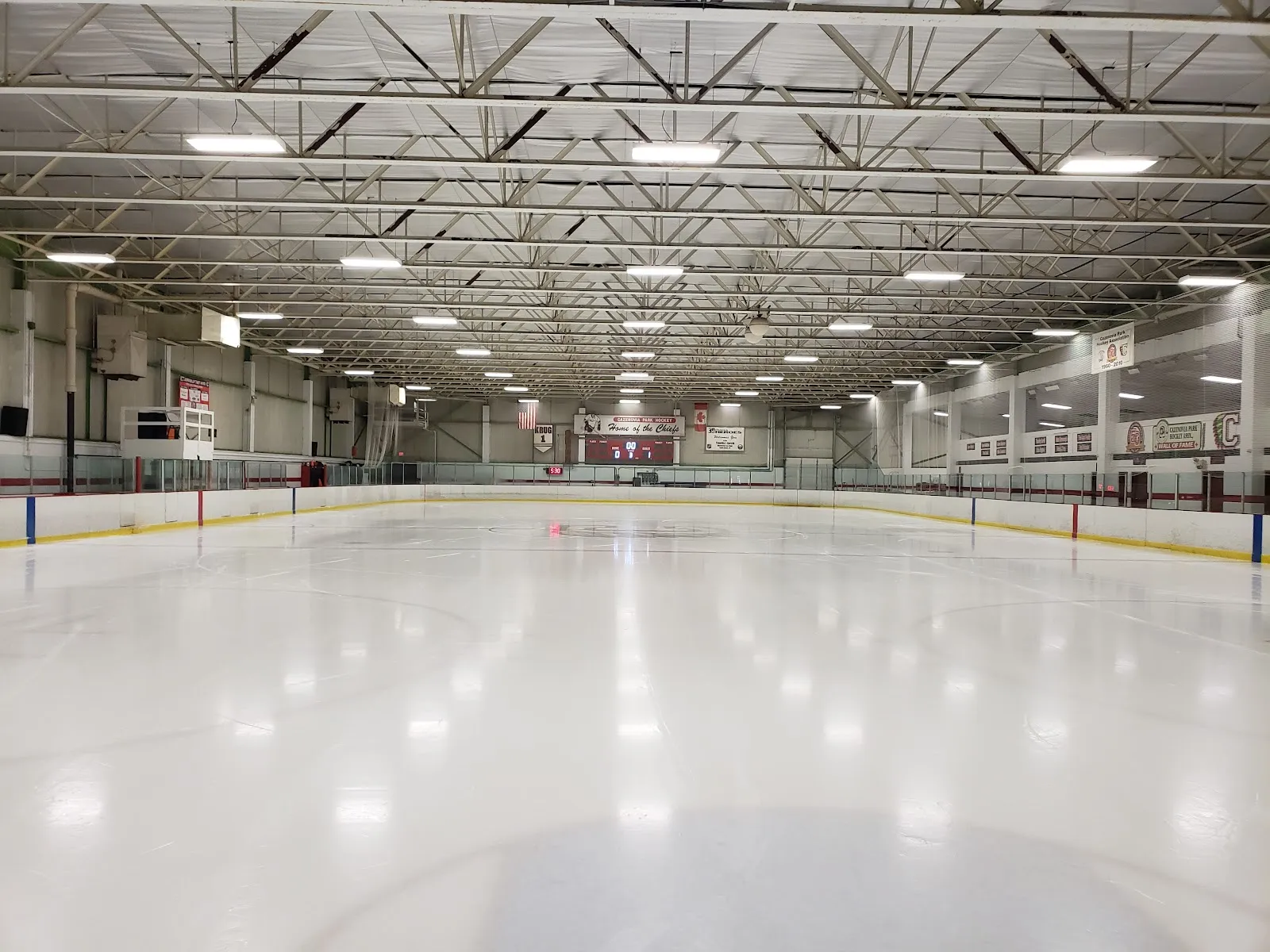Pista techada vacía vista desde el nivel del hielo hacia las vallas del fondo, con un marcador que dice "Cazenovia Park Hockey / Home of the Chiefs" y mantas de campeonato visibles, sin patinadores — Cazenovia Ice Rink